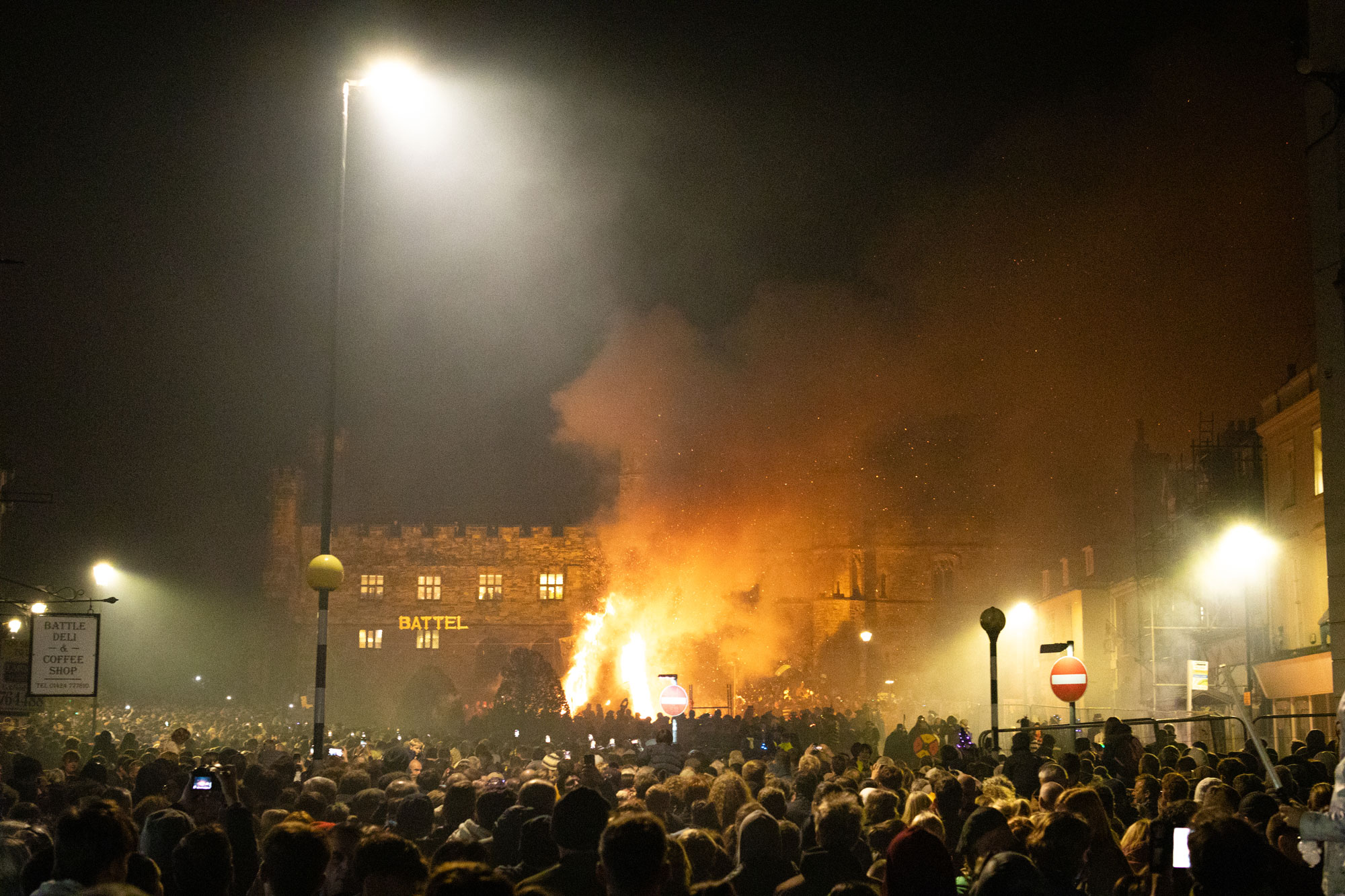 Eine Menschenmenge in Battle, England, die auf einem Platz ein großes Lagerfeuer beobachtet. Die Luft ist mit Rauch gefüllt und durch den Dunst sind verschiedene Straßenschilder sichtbar.