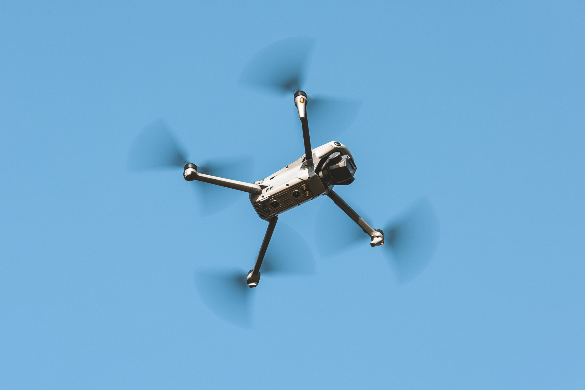 DJI Mavic 3 drone flying overhead against a clear blue sky, with propellers in motion.