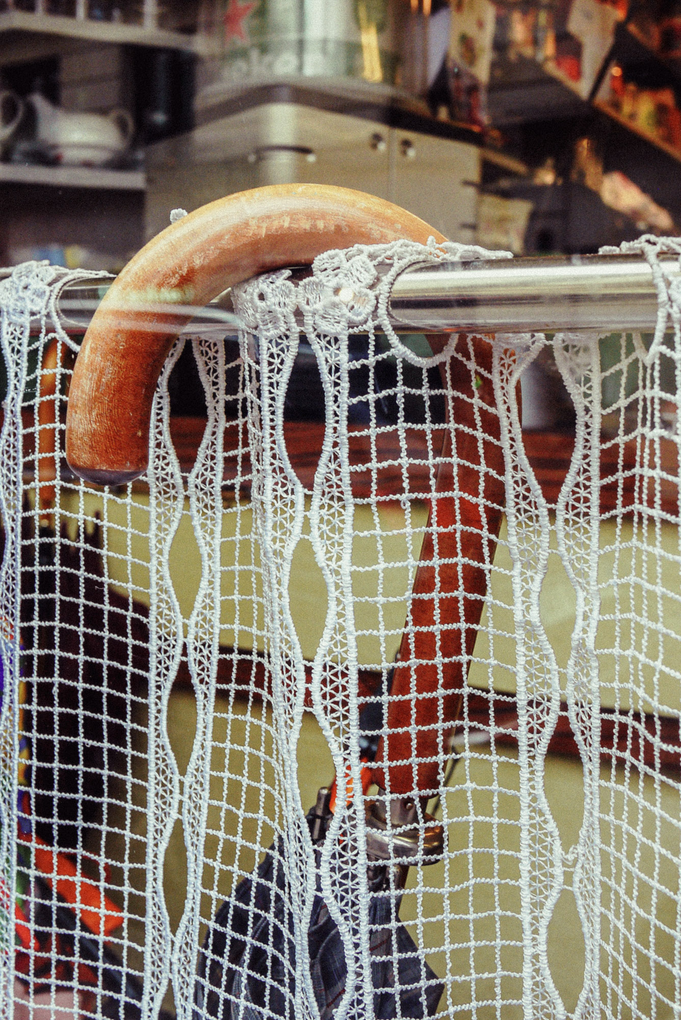 Nahaufnahme eines hölzernen Regenschirmgriffs, der hinter einem Spitzenvorhang über einer Vorhangstange im Fenster eines Pariser Cafés hängt. Durch den Stoff ist der Innenraum des Cafés zu sehen. Aufgenommen von Wesley Verhoeve mit einer Panasonic Lumix GF1.