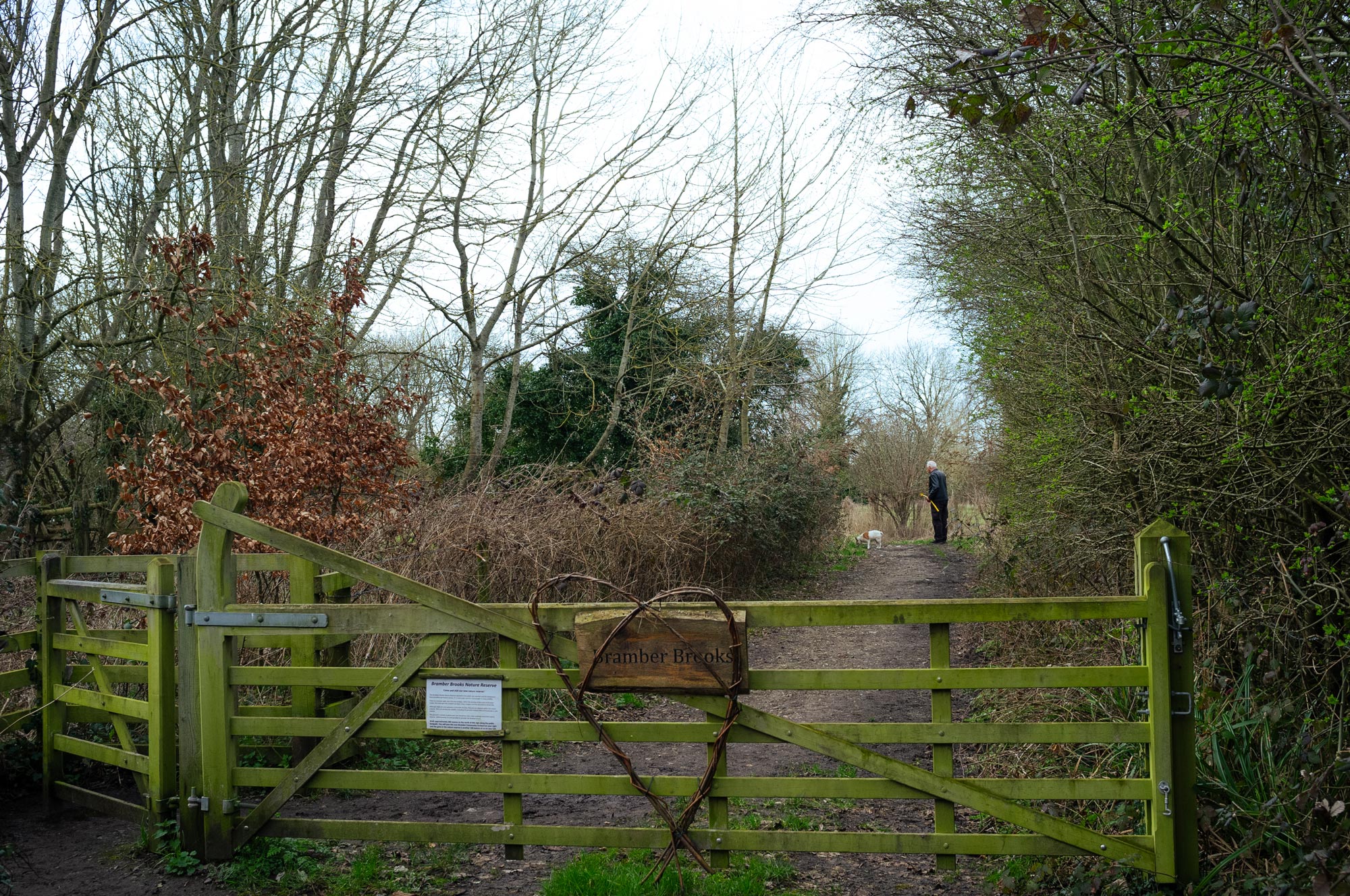 Een bewerkte foto van een persoon die hun hond uitlaat voorbij een gesloten houten poort met een bord waarop 'Bramber Brooks' staat en een hart gemaakt van wilgentenen