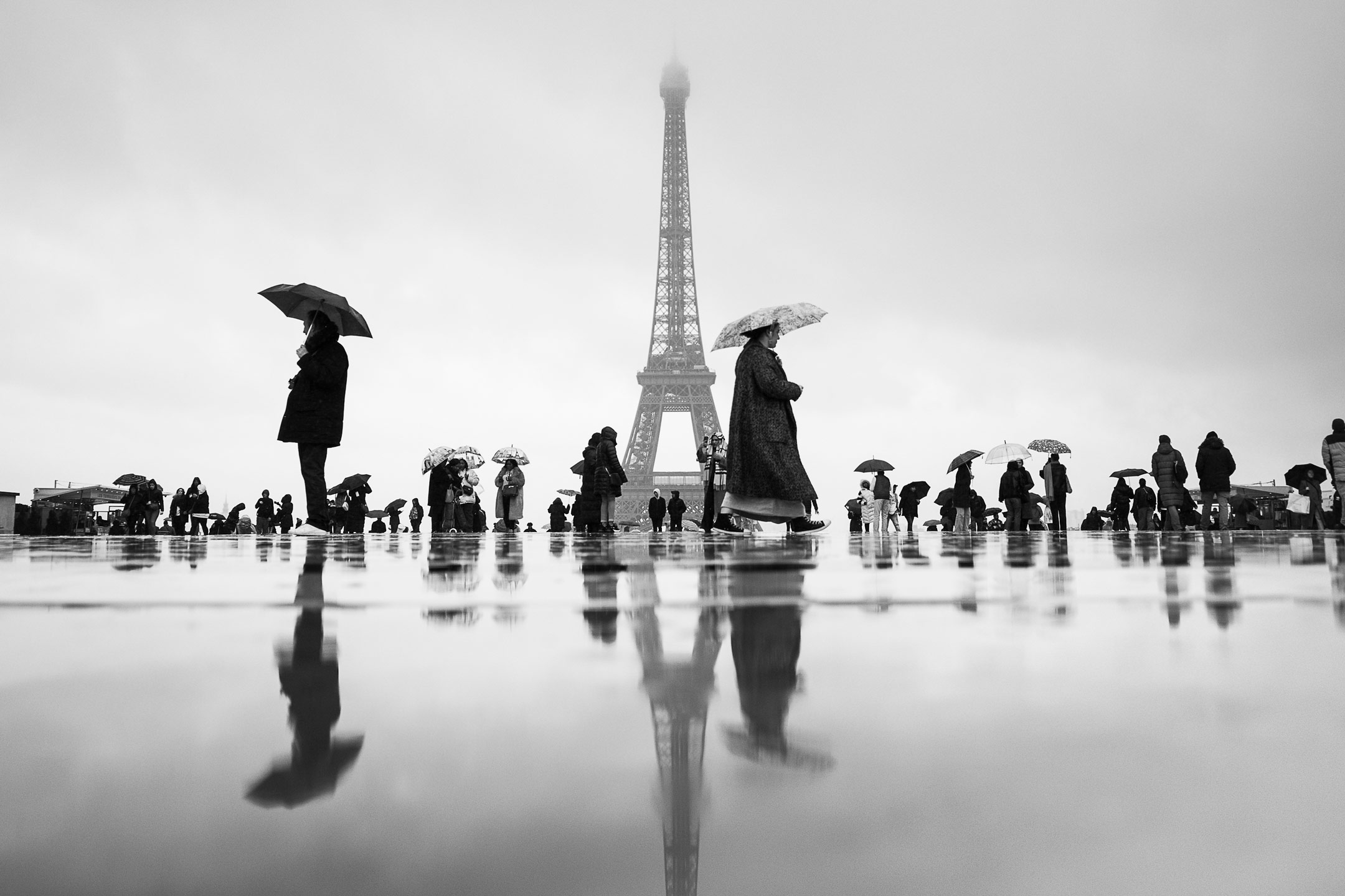 Black and white view of the Eiffel Tower in Paris with pedestrians holding umbrellas, reflected on wet pavement during a rainy day. Minimalist urban scene.