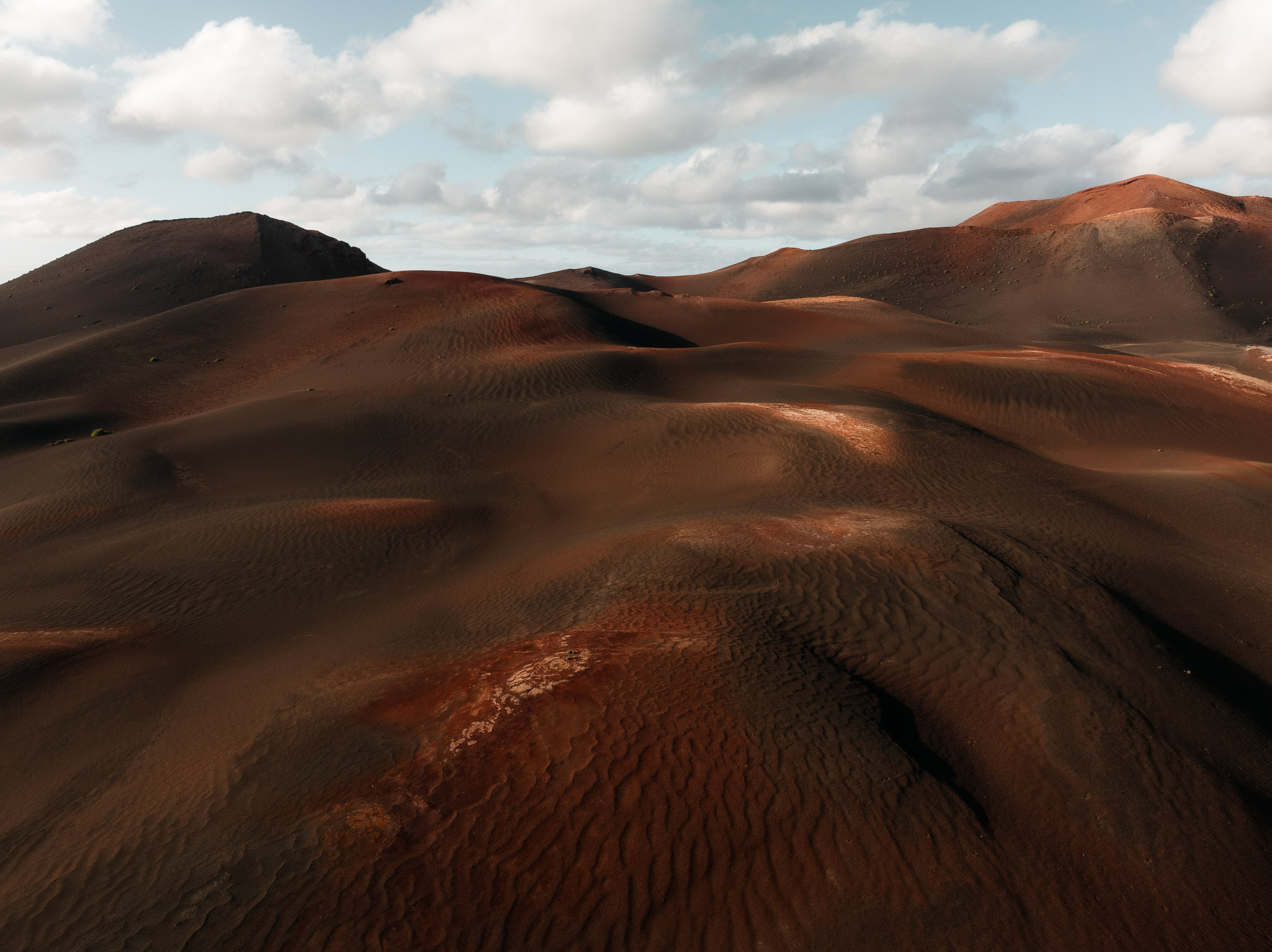 Aerial drone view of rolling dark desert dunes in Lanzarote with textured sand patterns under a partly cloudy sky.