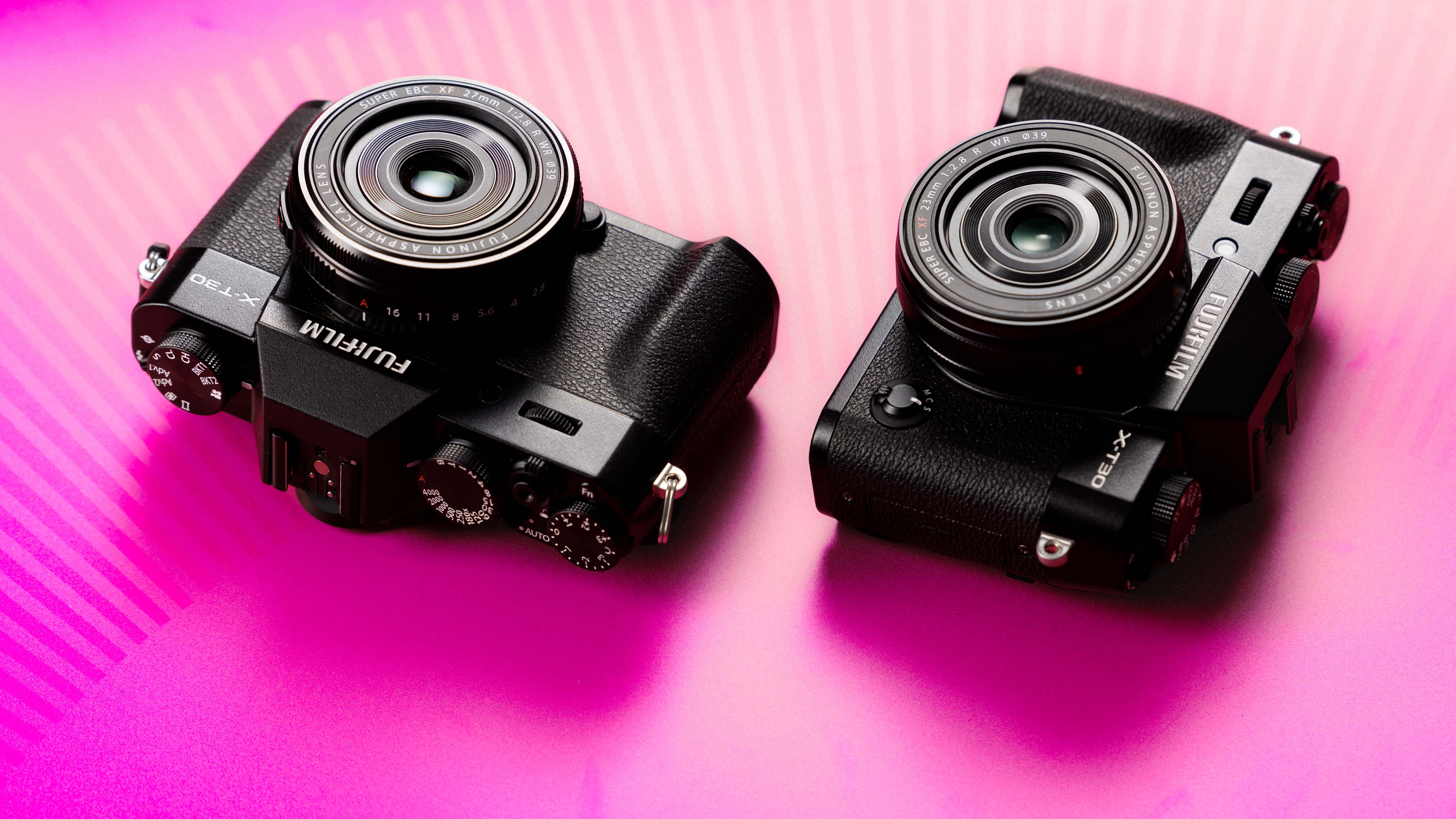 Two Fujifilm X-T30 mirrorless cameras with Fujinon lenses displayed on a vibrant pink background, showcasing compact design and photography gear in a studio setup.