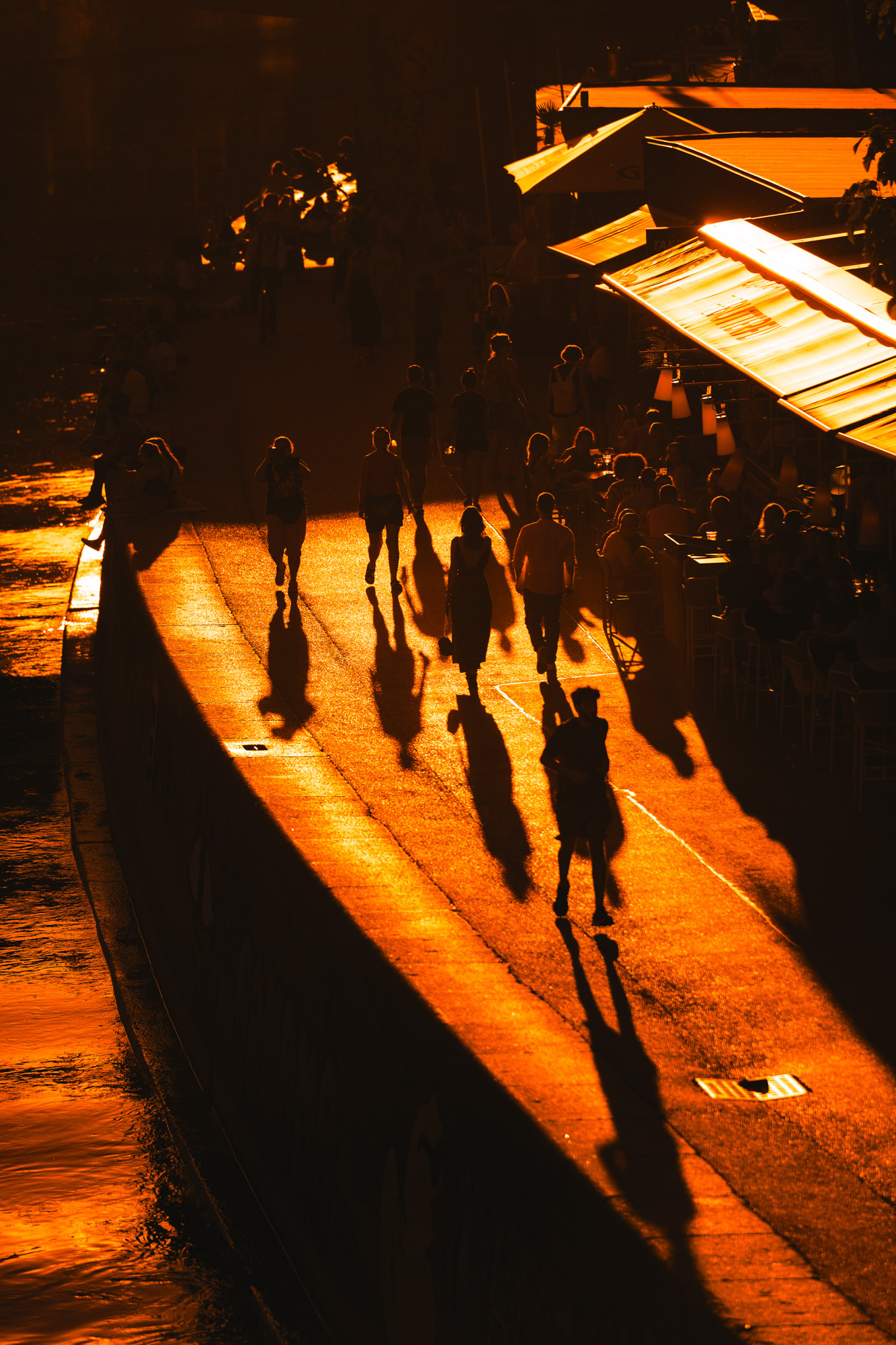 Spaziergänger:innen als dunkle Silhouetten am Donaukanal bei tief stehender Sonne. Lange Schatten ziehen über den Weg, rechts Terrassen im Licht.