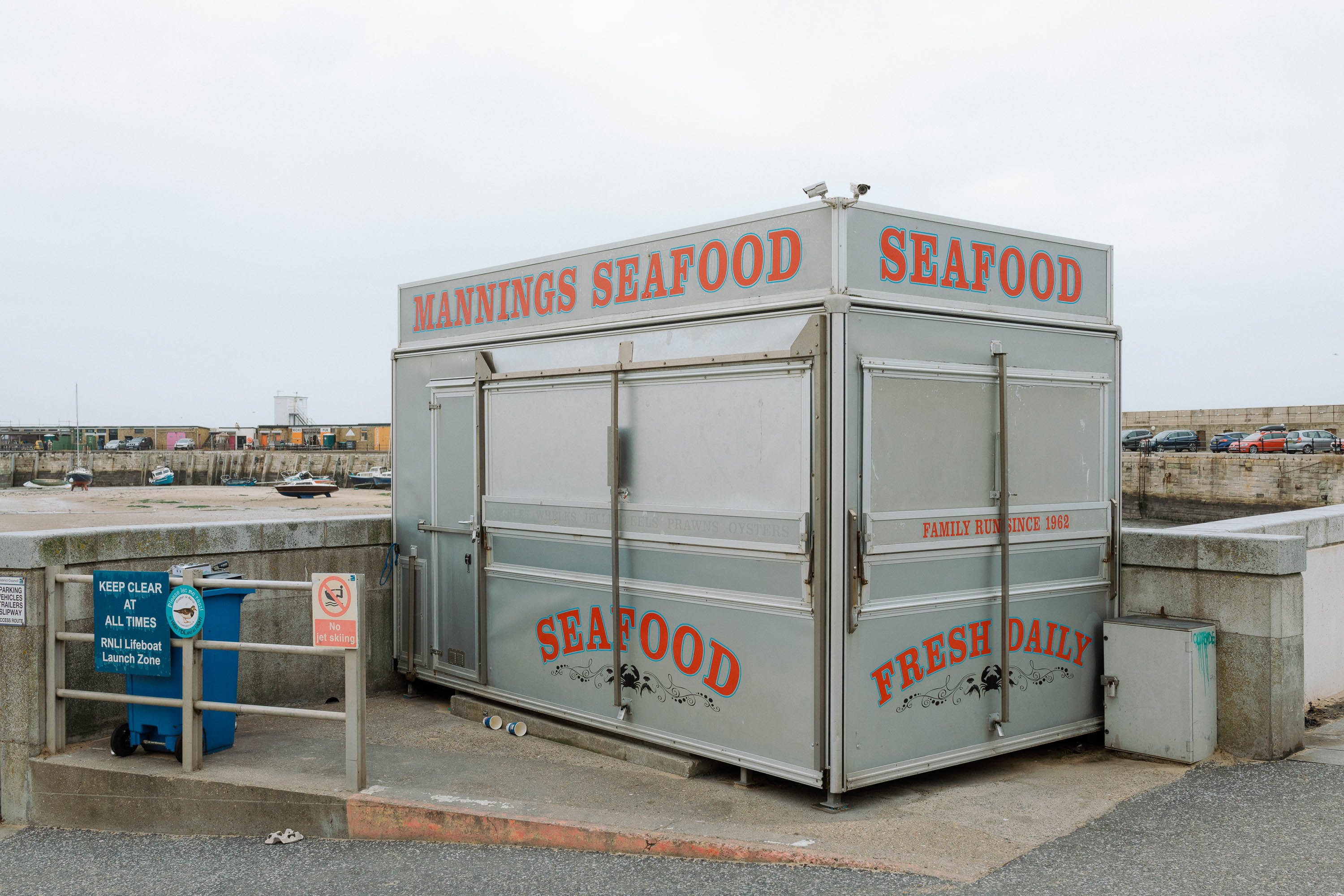 Ein geschlossener Fisch- und Meeresfrüchte-Kiosk mit der Aufschrift „Mannings Seafood“, fotografiert auf einem Hafensteg mit Booten, Steinmauern und einem bewölkten Himmel im Hintergrund. Aufgenommen von Connor Redmond mit einer Sony RX1R III.