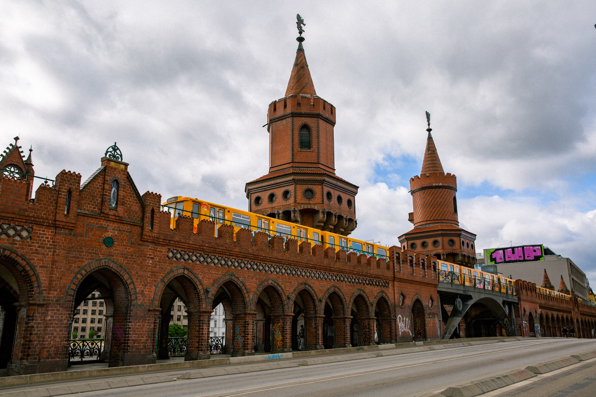 Yellow U-Bahn train crossing Berlin’s Oberbaum Bridge, with red-brick arches and towers beneath a dramatic cloudy sky. Shot by Austin Fassino.