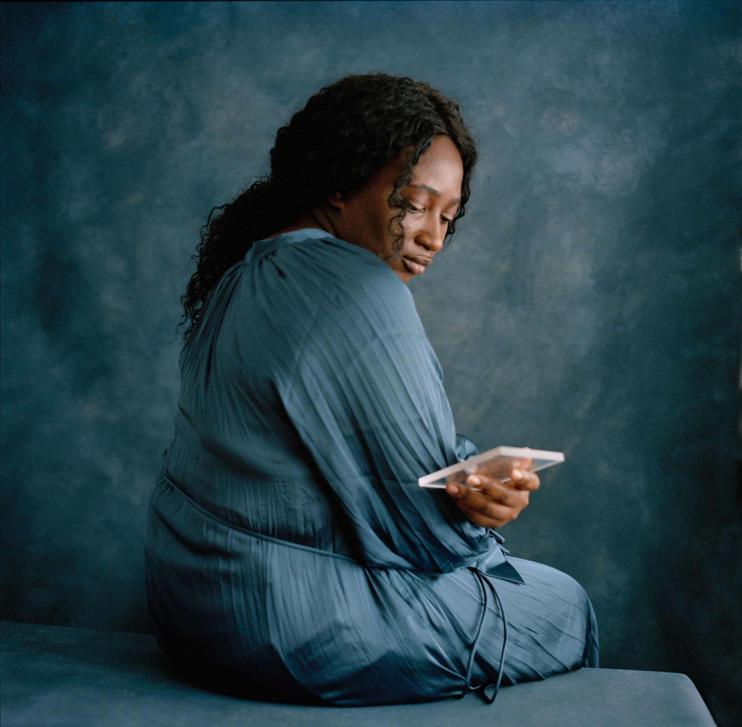 Black woman in blue clothes sitting on a blue bench looking at herself in a piece of glass she is holding, shot by Nicole Benewaah