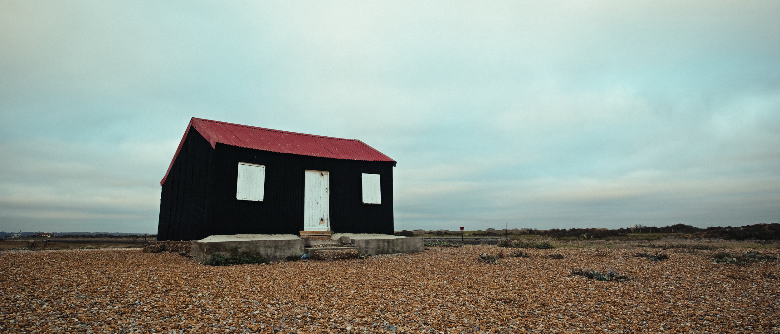 Ein Farbfoto von einer rot-schwarzen Strandhütte, umgeben von einem Kiesstrand im Rye Harbour Nature Reserve, aufgenommen mit einer Sigma DP0 Quattro von Jakub Golis