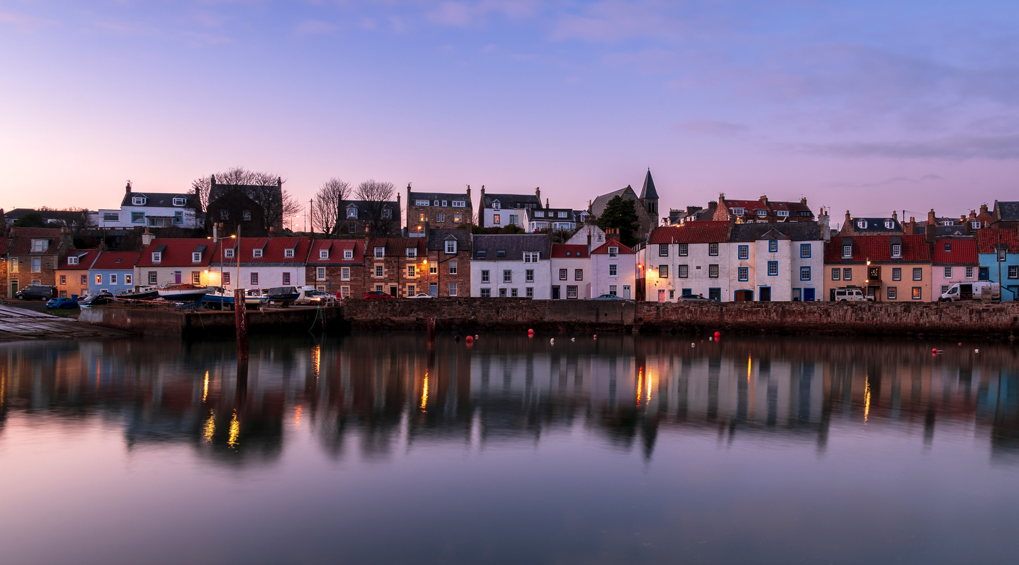 Case colorate di un villaggio costiero al tramonto, con luci accese riflesse nell'acqua calma del porto.