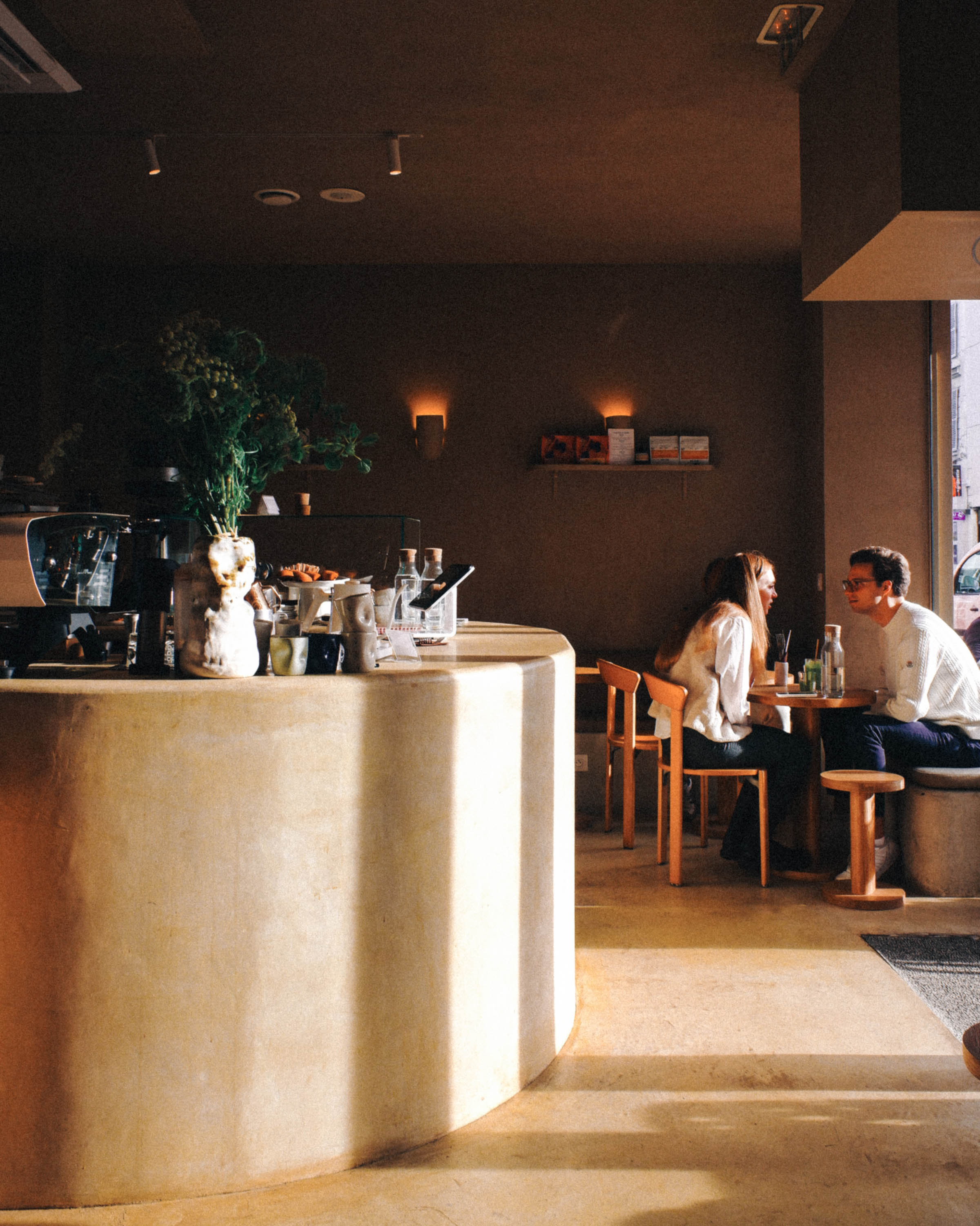 A calm Parisian café moment with two people chatting at a wooden table, framed by warm sunbeams, soft shadows and a clean, contemporary interior. Shot by Wesley Verhoeve on a Fujifilm XF10.