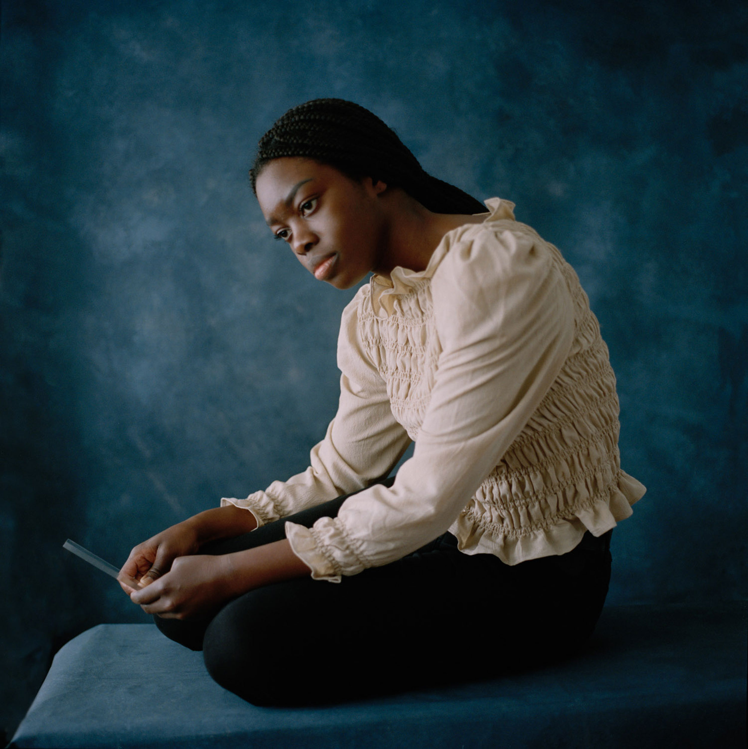 Black woman sitting and looking into the void, in front of a blue background, shot by Nicole Benewaah