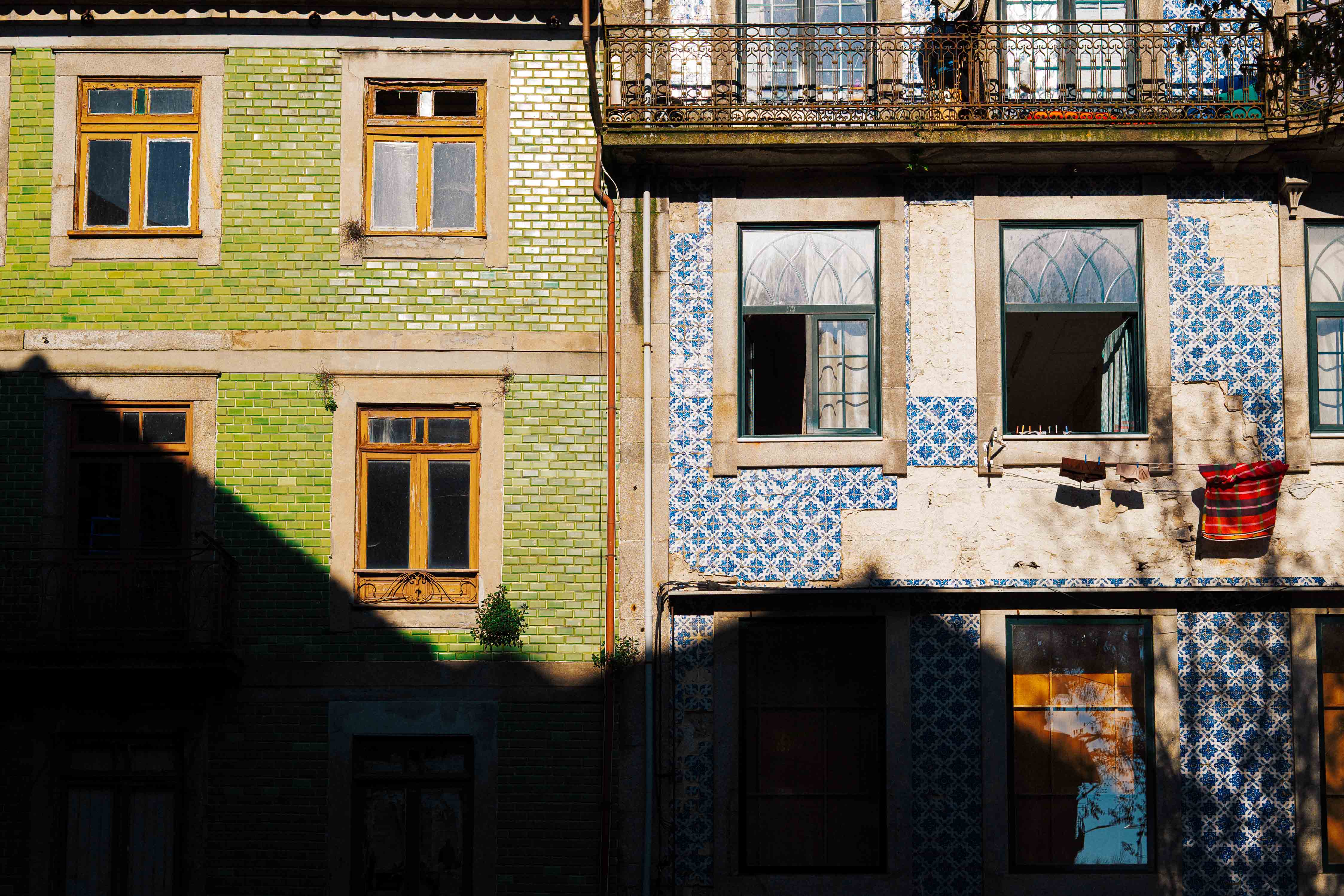 Contrasting green and blue azulejo-tiled façades in Porto, with sunlight and shadow dividing the buildings across their windows.
