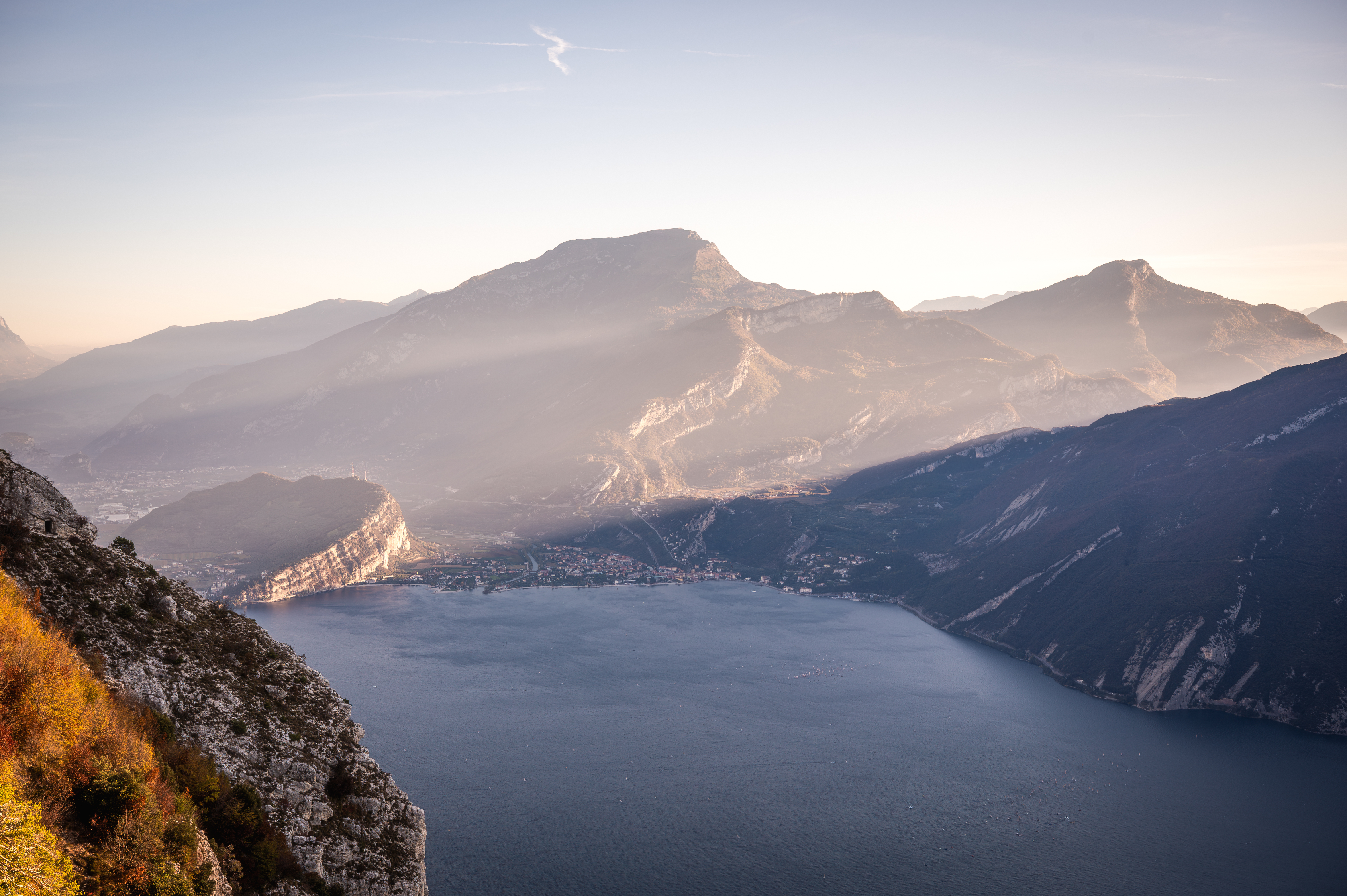 Vista dall'alto sul Lago di Garda al tramonto, con montagne, foschia e una cittadina sulla riva.