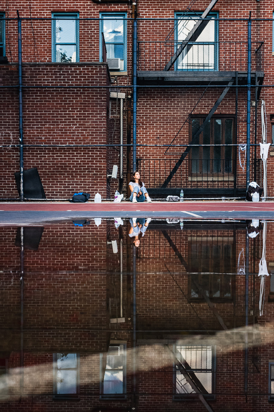 Persoon zittend op basketbalveld met plas en weerspiegeling met bakstenen gebouw van New York City op de achtergrond, gefotografeerd door Manuel Pena.