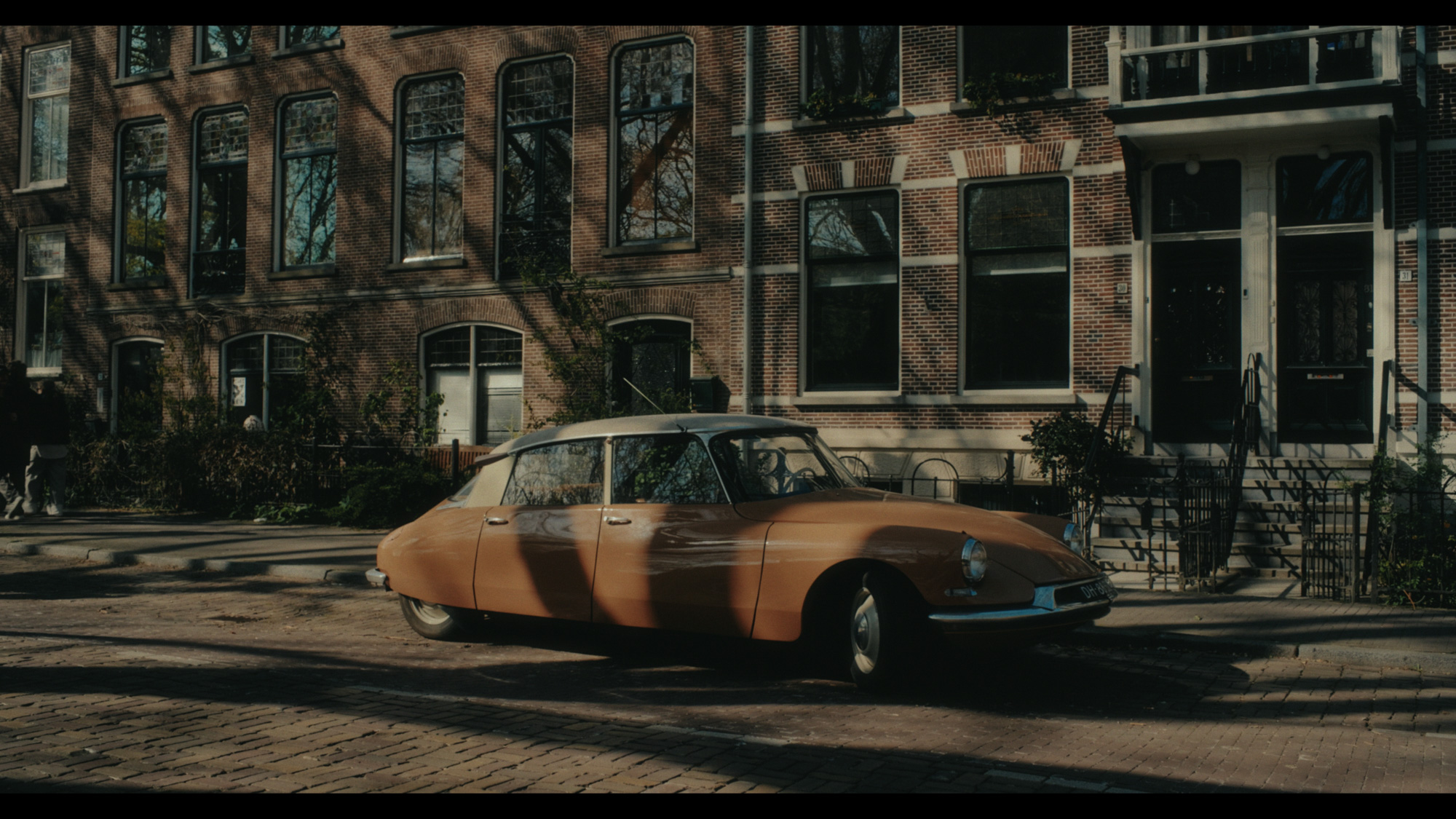 An orange vintage car is parked along a brick street in front of traditional multi-story residential buildings with large windows.