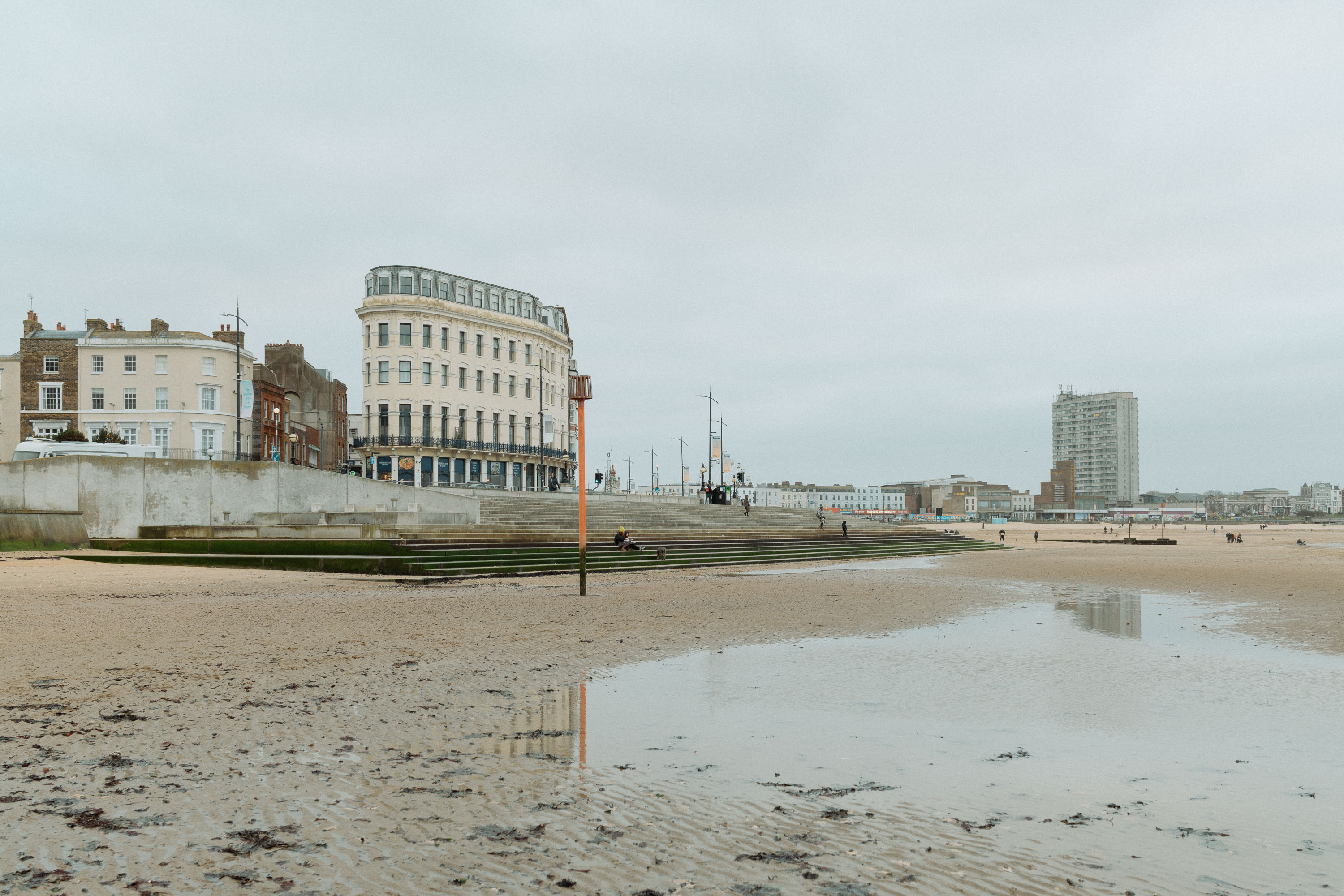 A wide view of a Margate seaside promenade and beach at low tide, with historic buildings, steps leading to the sand and reflections in shallow pools under an overcast sky. Photo by Connor Redmond with the Sony RX1R III.
