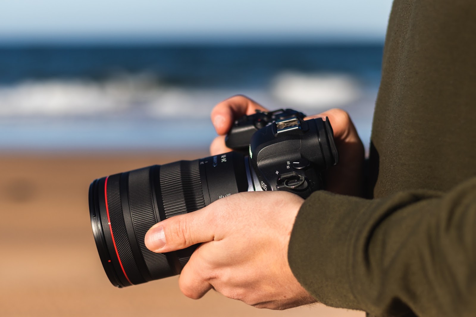 A close-up of a person’s hands and arms, wearing a dark green jumper, holding a Canon DSLR and lens, with an out-of-focus beach in the background, shot by Jonas Hanspach
