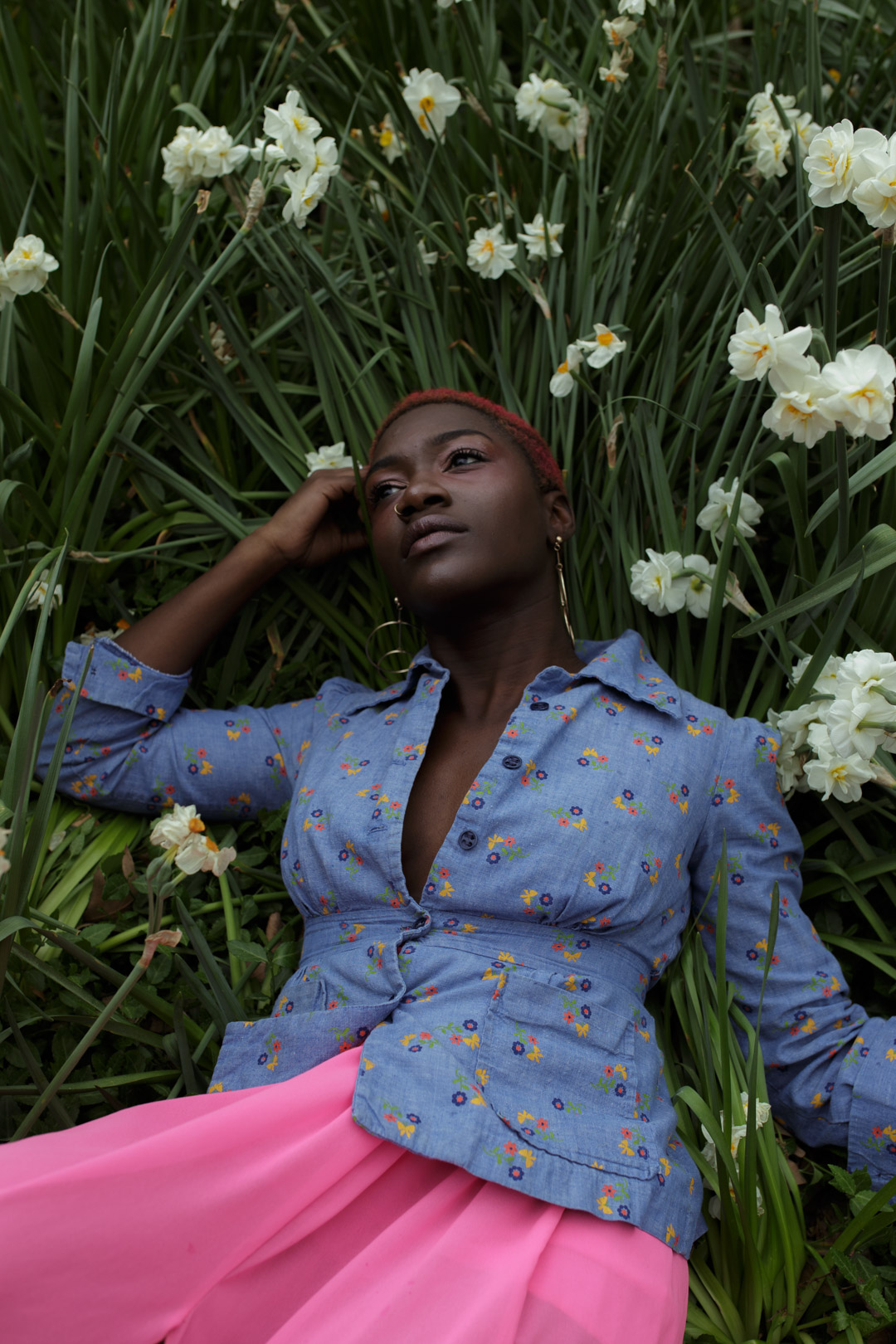 Photo of black woman laying in grass with a pink skirt and blue shirt surrounded by flowers