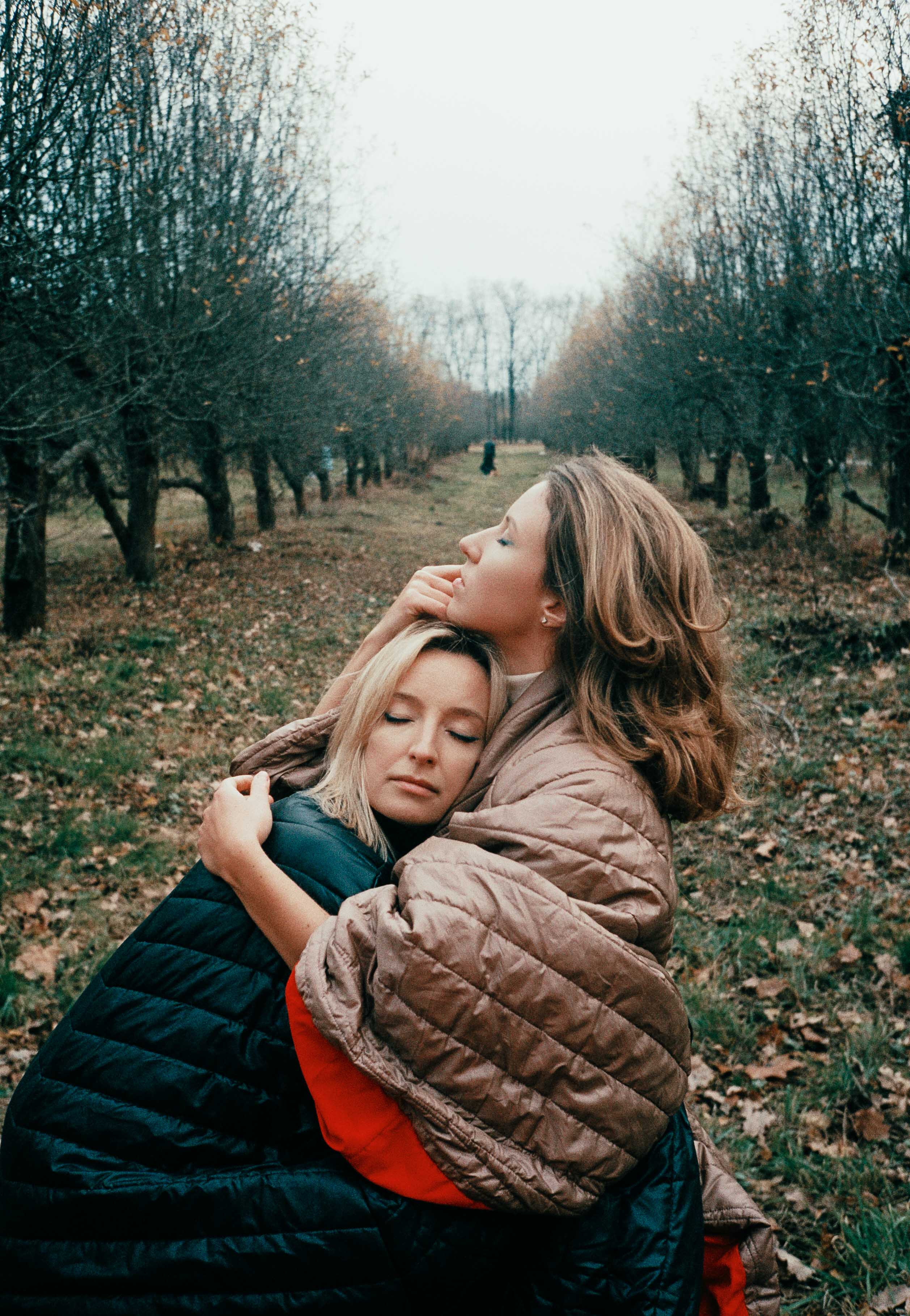 Emotional autumn scene of two people wrapped in cozy jackets, embracing between rows of leafless trees. Shot by Alina Rudya on ISO 400 film.