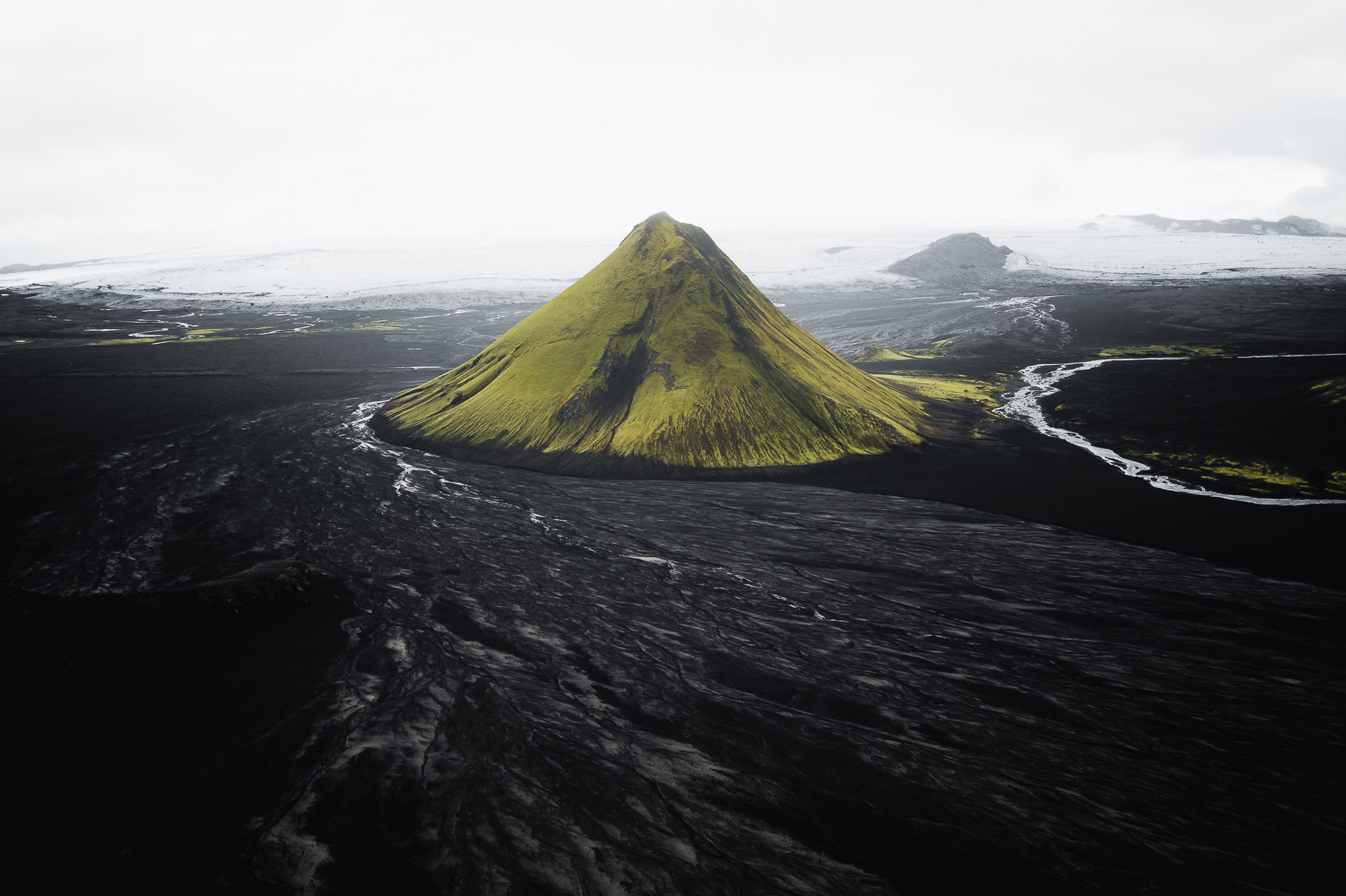 An aerial drone landscape photo of a large green hill surrounded by grey in Iceland, with snow and mountains in the background, shot by Paul Watson with a DJI Mavic 2 Pro