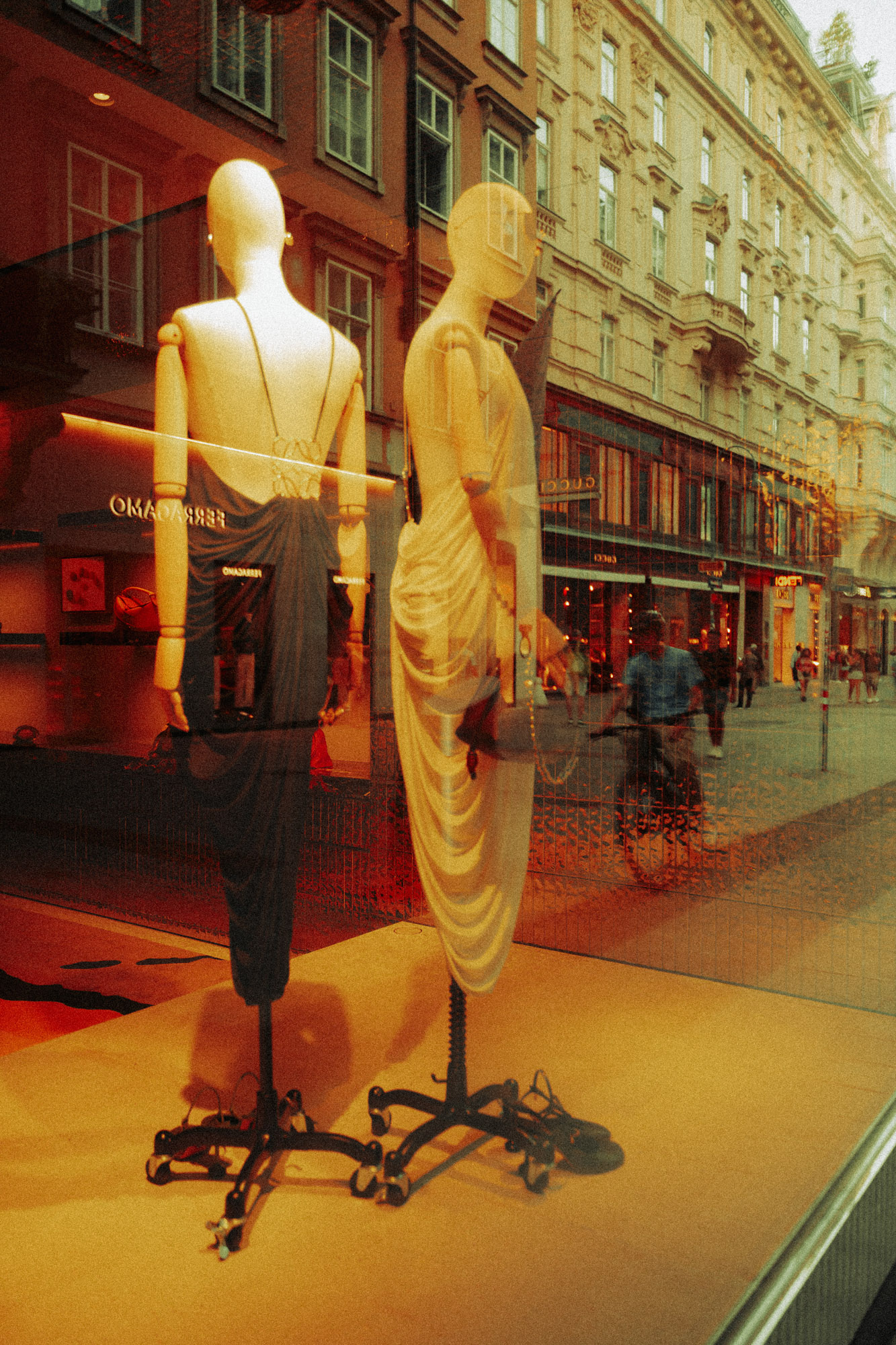 Farbfoto eines Schaufensters in Wien mit zwei Schaufensterpuppen in eleganter Kleidung. In der Scheibe spiegeln sich eine Straße, Passant:innen sowie historische Fassaden, wodurch ein vielschichtiger, dokumentarischer Eindruck entsteht. Aufgenommen von Dhan Limwattana mit einer Nikon Z5 II.
