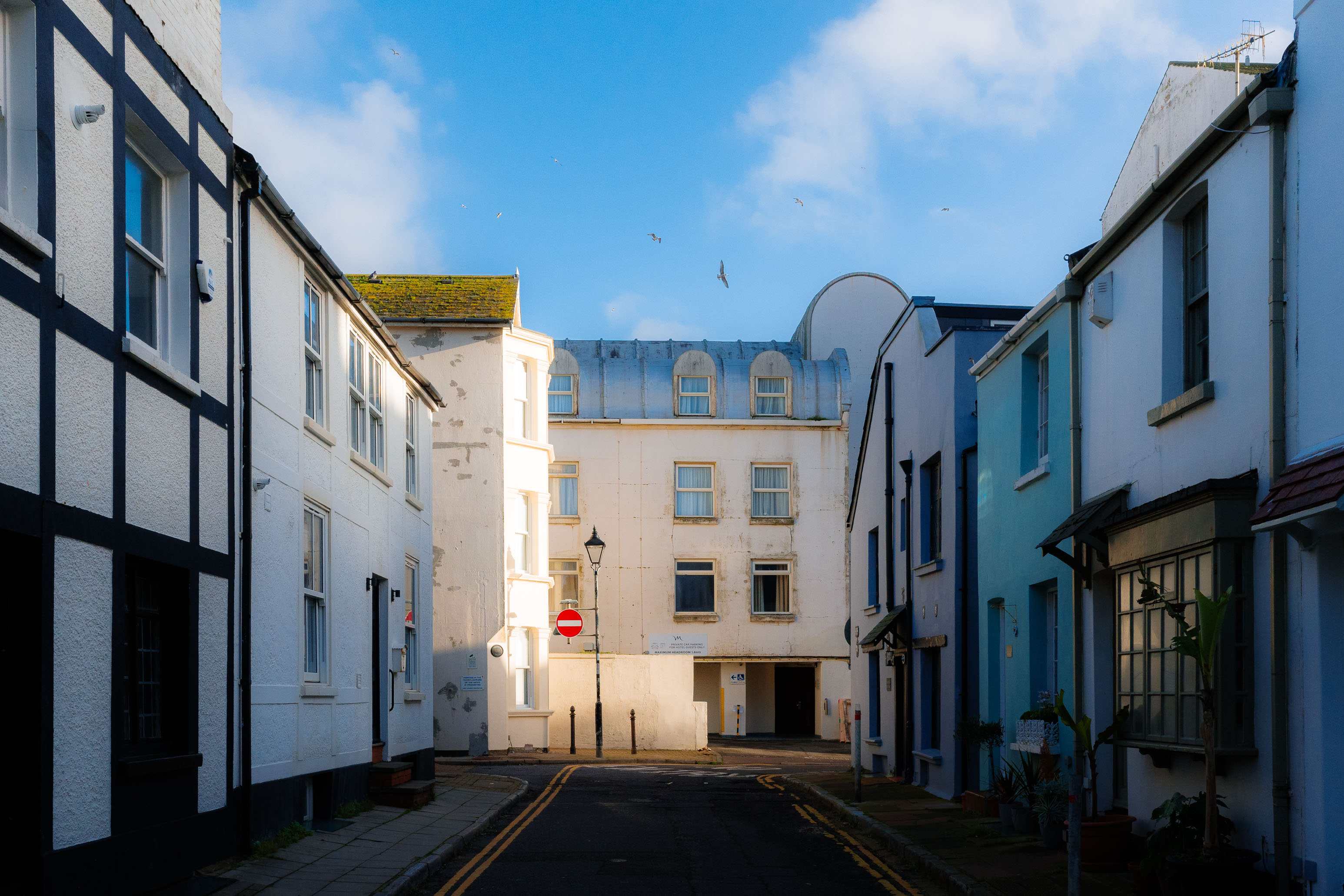 Eine ruhige Streetaufnahme zwischen weißen und pastellfarbenen Gebäuden bei natürlichem Tageslicht. Im Hintergrund, sanftes Sonnenlicht und klarer blauer Himmel. Aufgenommen mit der Ricoh GR IV.