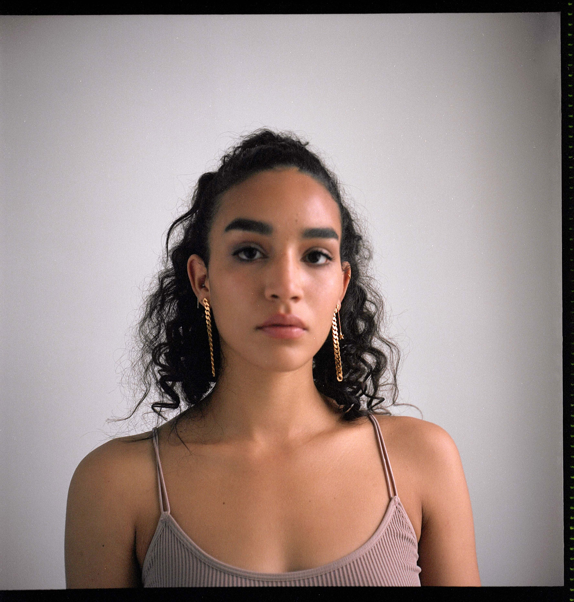 Portrait of a Person of Color with long gold earrings looking straight into the camera with a serious look, against a grey background, shot by Samia Rachel.
