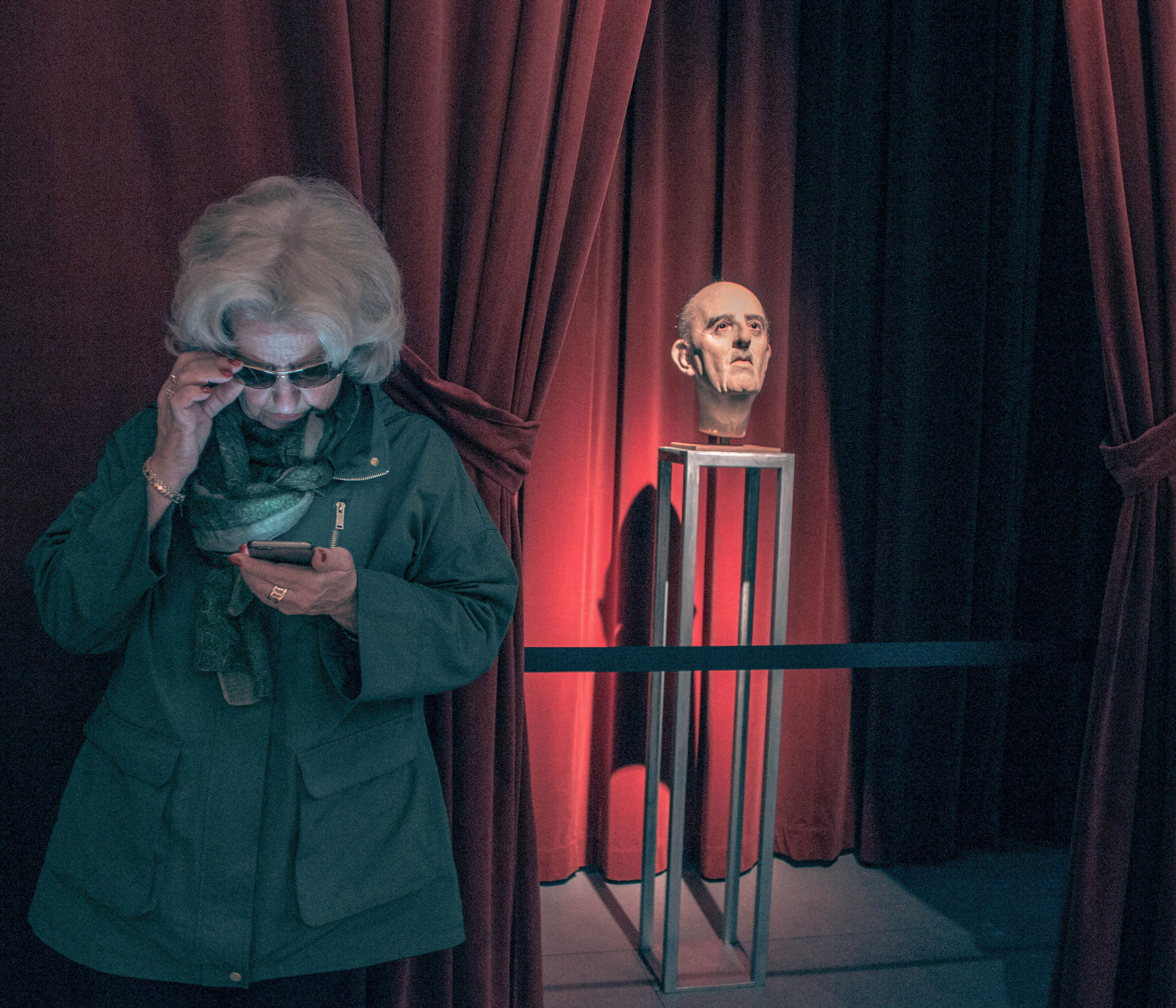A person with white hair wearing a dark green jacket looks down at their phone in front of a waxwork bust of Spanish dictator Francisco Franco surrounded by red curtains, shot by Jordi Jon Pardo with a Canon EOS 700D