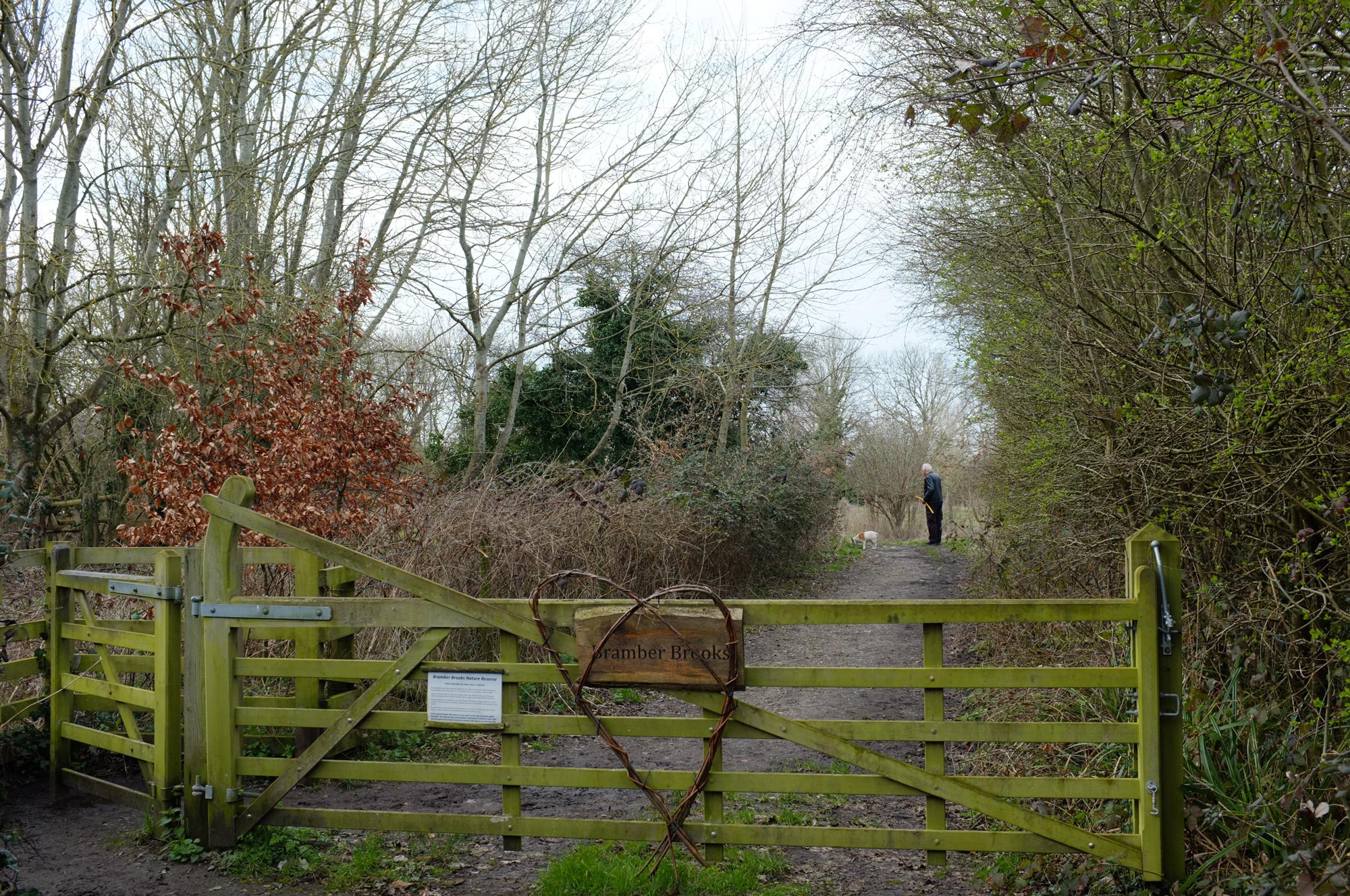 Unedited photo of a person walking their dog beyond a closed wooden gate with a sign that reads ‘Bramber Brooks’ and a heart made out of wicker