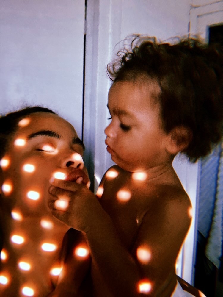 Un parent et son enfant éclairés par une lumière vive passant à travers les interstices d'un rideau, prise de vue par Charlotte Yonga.