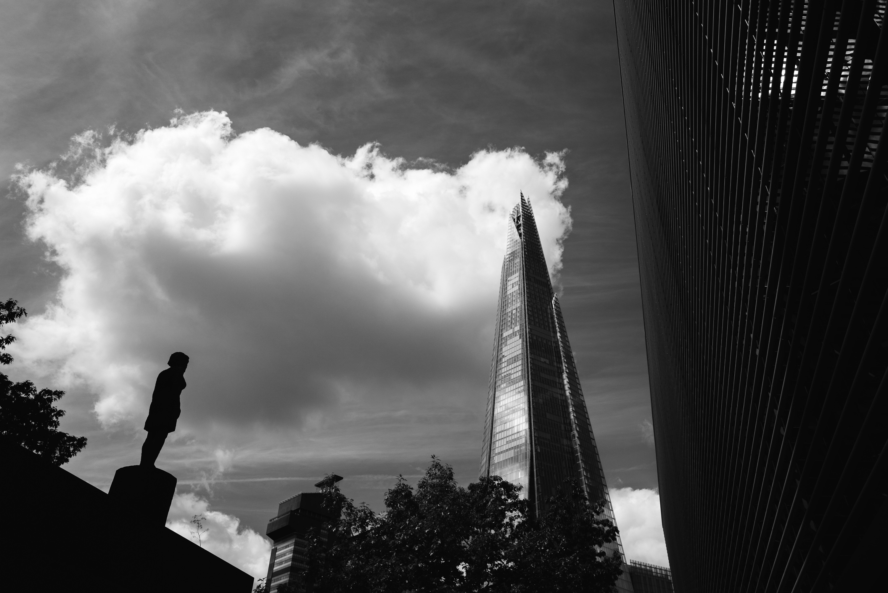 Une photo en noir et blanc du Shard à Londres. Il y a un bâtiment d'un côté et une statue de l'autre, ainsi qu'un gros nuage remplissant la majeure partie du ciel. Photo prise avec un Leica Q2