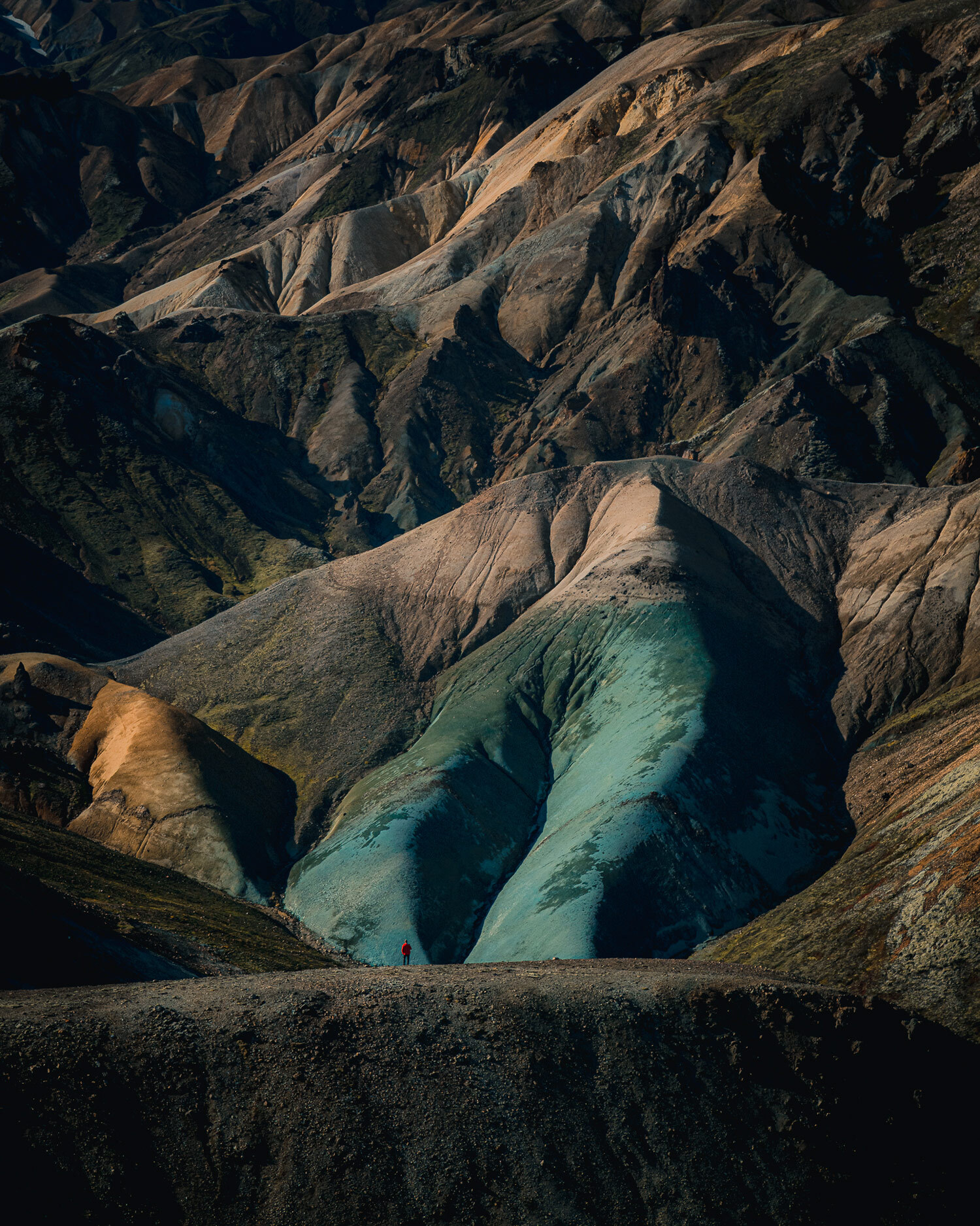 A figure in a red coat stands on top of a ridge in a mountain range in Iceland, one side of a mountain has a distinctive light-blue colour, shot by Paul Watson with a Panasonic S1R
