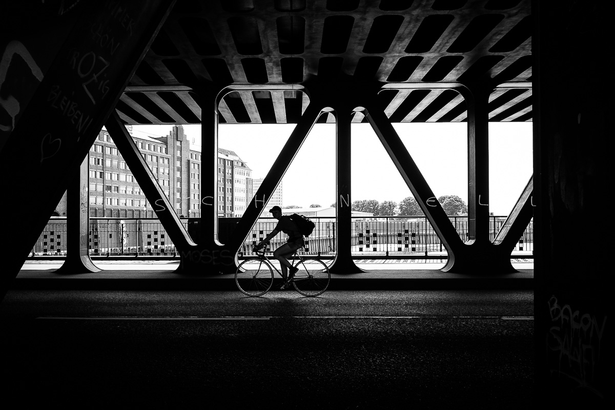 The silhouette of a cyclist on a covered iron bridge with cityscape in background, black-and-white street photography by Marco Larousse