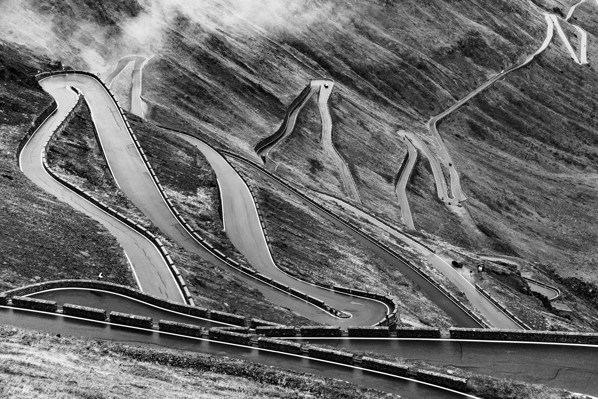 Schwarz-Weiß-Foto der atemberaubende Kurven eines legendären Bergpasses. Die gewundenen Straßen bilden ein faszinierendes Muster in der rauen Landschaft und machen dieses Bild zu einer perfekten Inspiration für Abenteuer-, Reise- und Kunstfotograf:innen.
