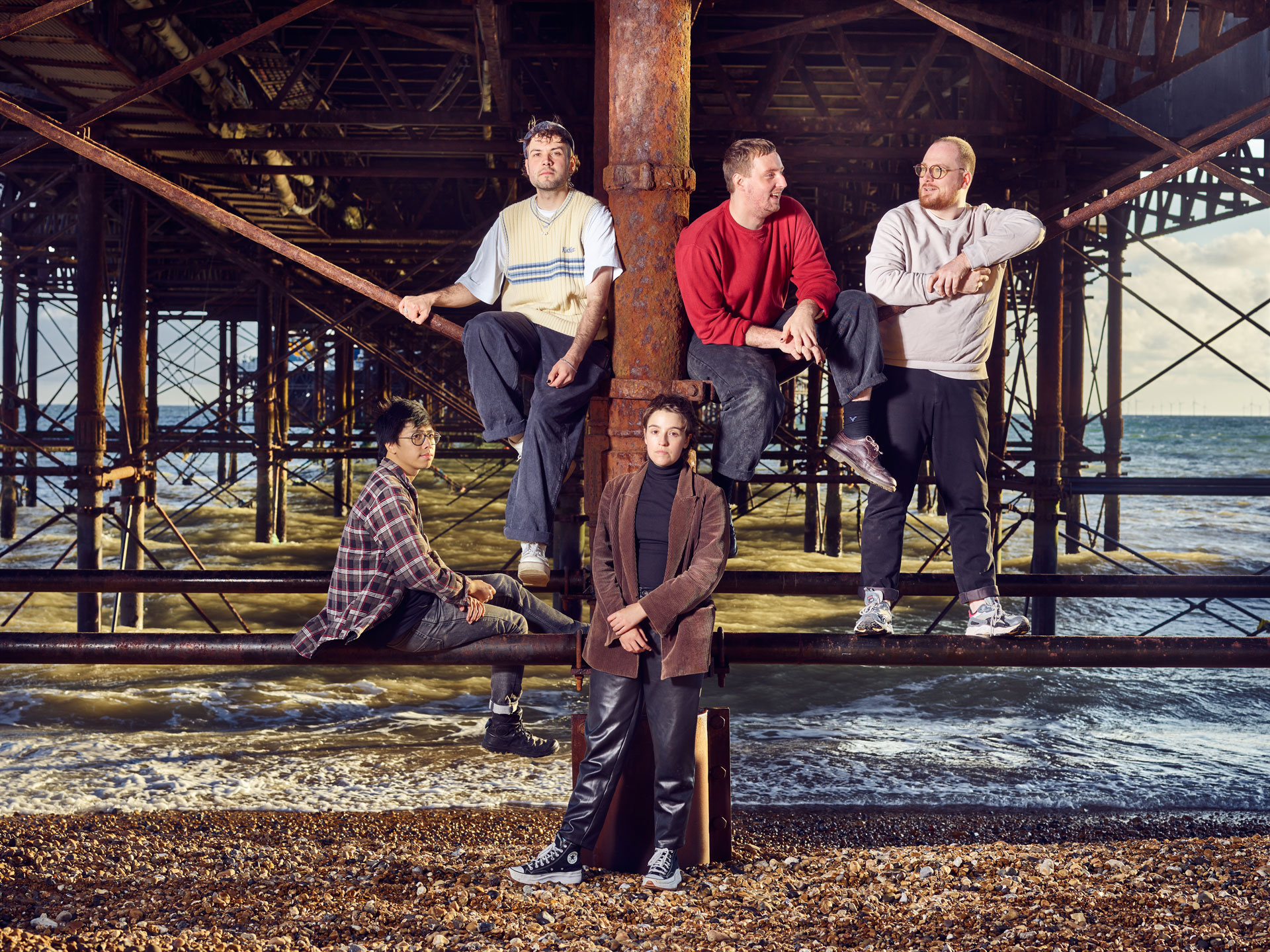 The band ‘Flirting’ sitting on the rusty supports under Brighton Palace Pier, with sea waves in the background, photo by Ki Price