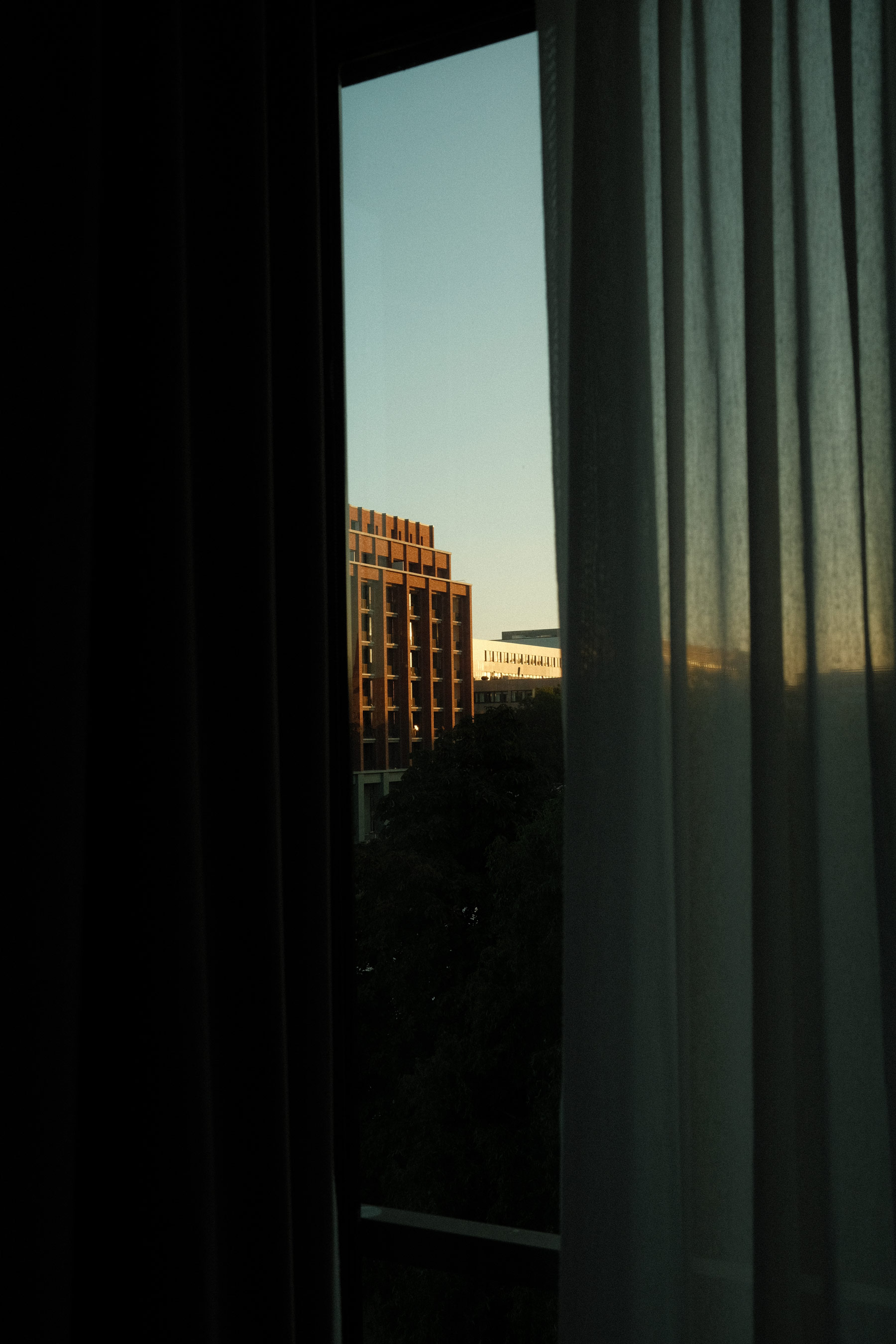 View from a partially curtained window looking out at a building, photo by Hermann Furin with a Fujifilm X100V