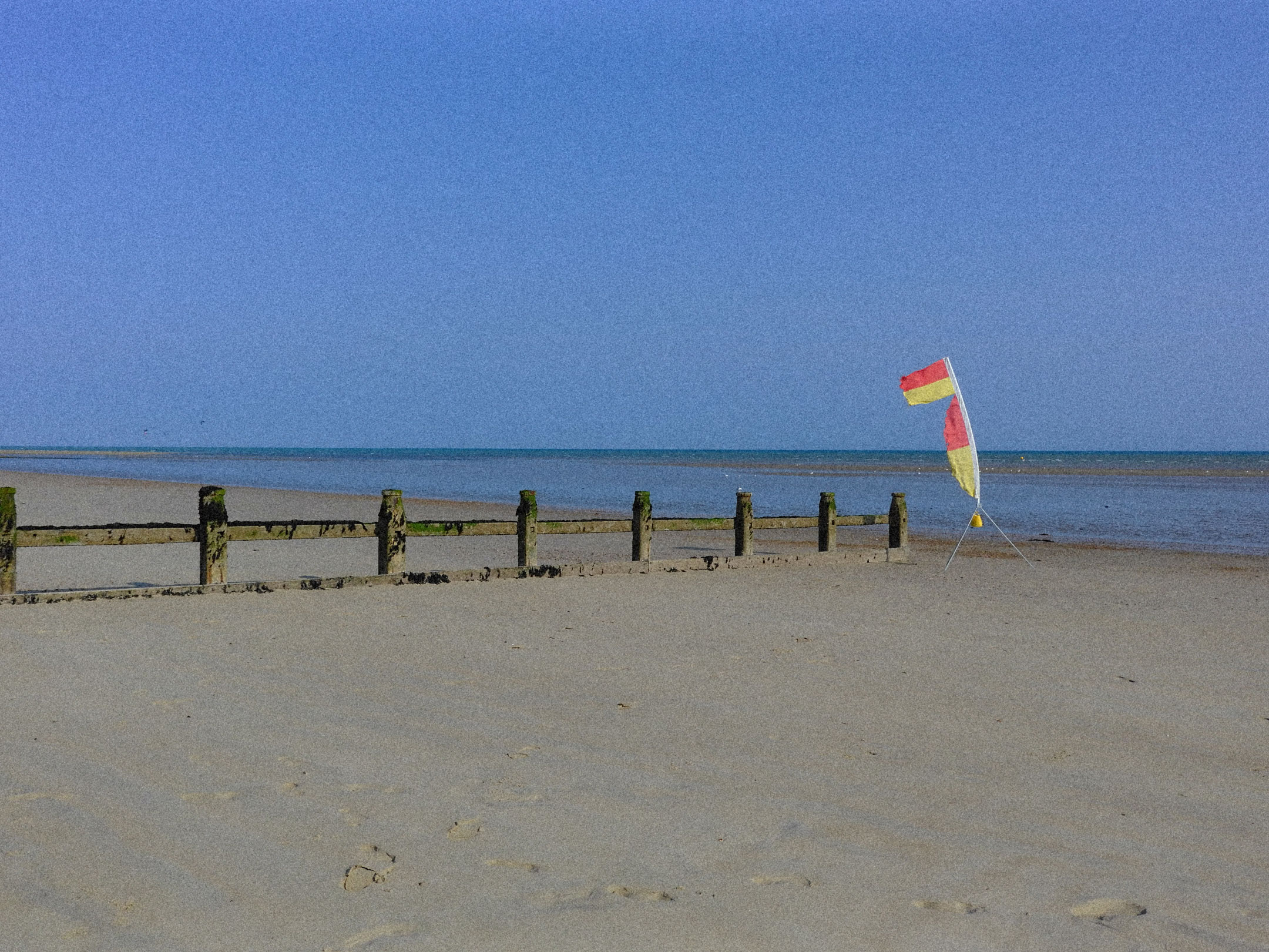 Eine rot-gelbe Strandflagge und hölzerne Buhnen im Sand bei Ebbe. Im Hintergrund, das Meer.