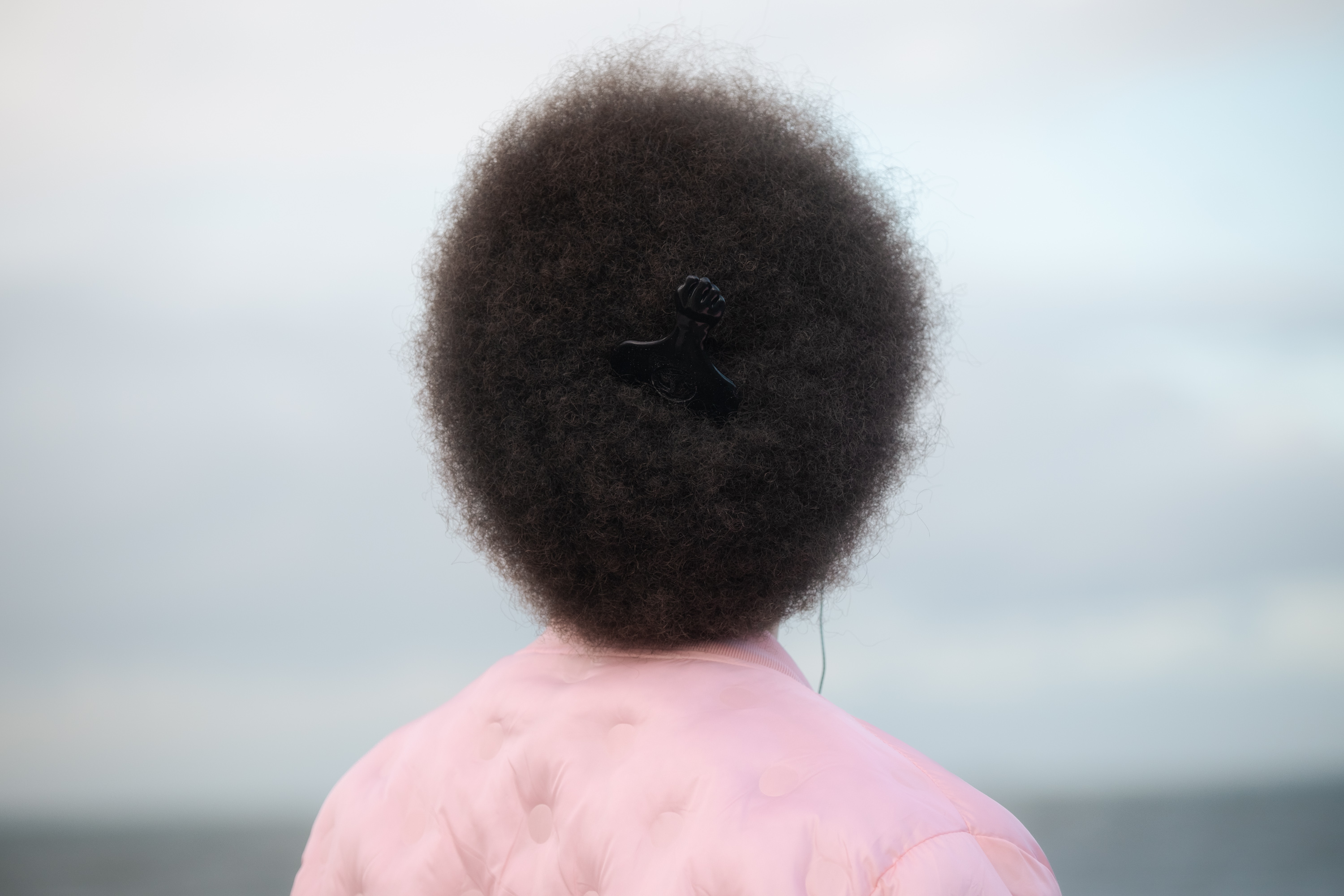 Aufnahme einer Person von hinten, die auf das Meer hinausschaut, eine rosa Jacke trägt und ihr Haar als Afro mit einem Afrokamm trägt, aufgenommen mit einer Fujifilm X-T5