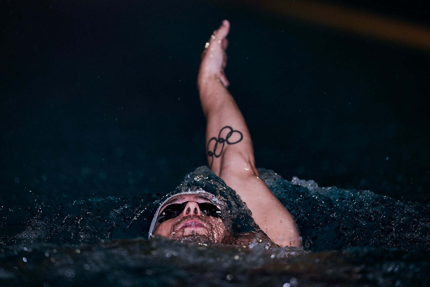 Een zwemmer die de rugslag doet met één arm met de Olympische ringen als tatoeage en zijn gezicht omhoog uit het water.
