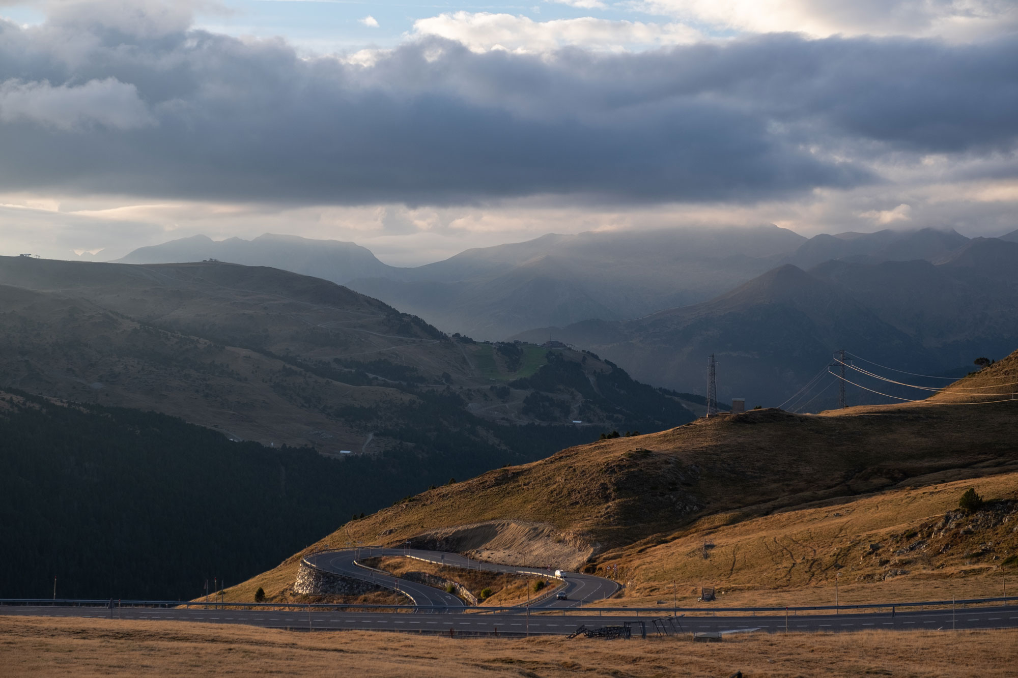 A mountainous landscape with winding roads. The foreground is clear and warm, illuminated by the setting sun's light, while the background is cloudy and appears colder.