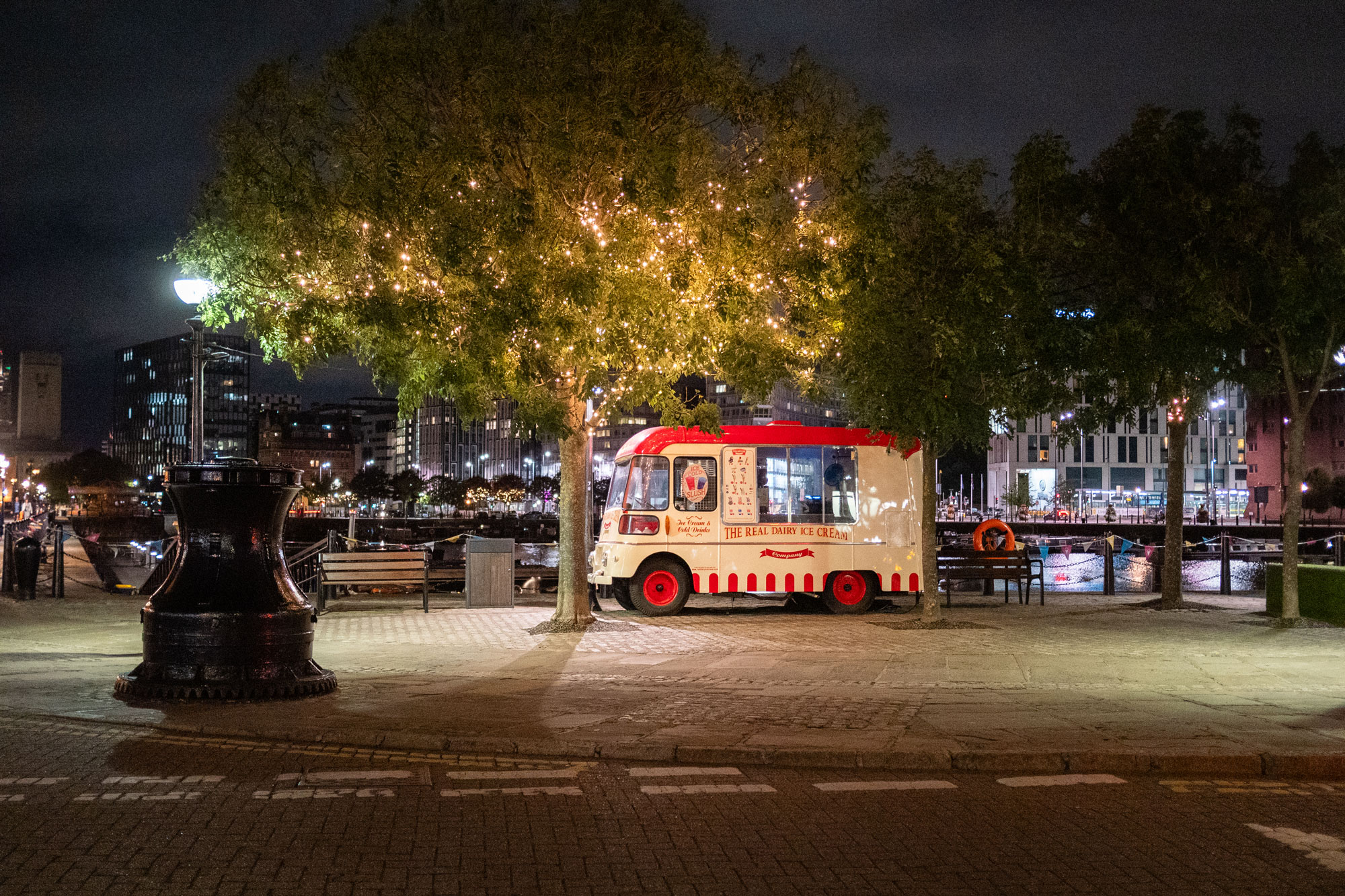 Ein rot-weißer Eiswagen im Dunkeln an einer Hafenpromenade. Davor ein Baum mit einer Lichterkette in der Krone.
