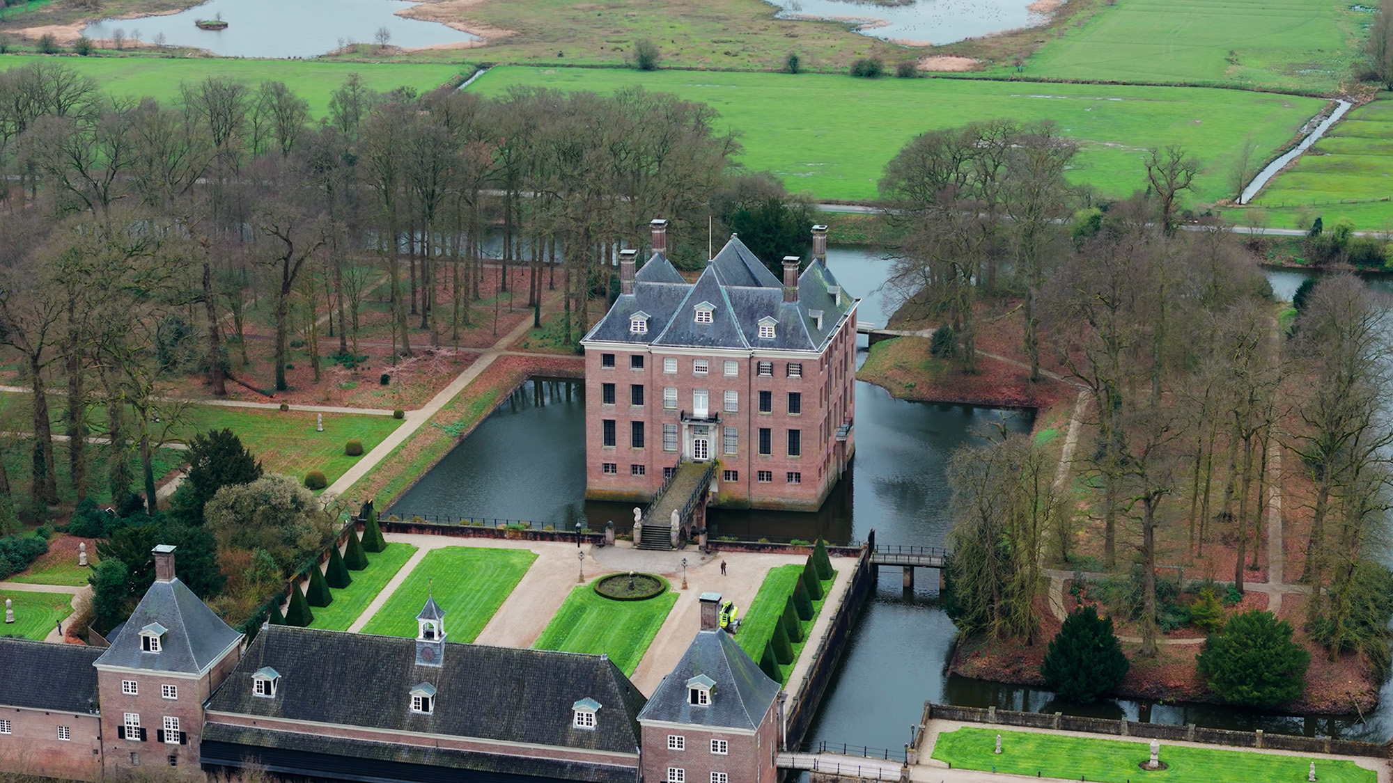 Een prachtig luchtbeeld van een historisch kasteel, omringd door brede grachten en symmetrische tuinen, gelegen in een serene groene omgeving, geschoten met de DJI Air 3, 70 mm.