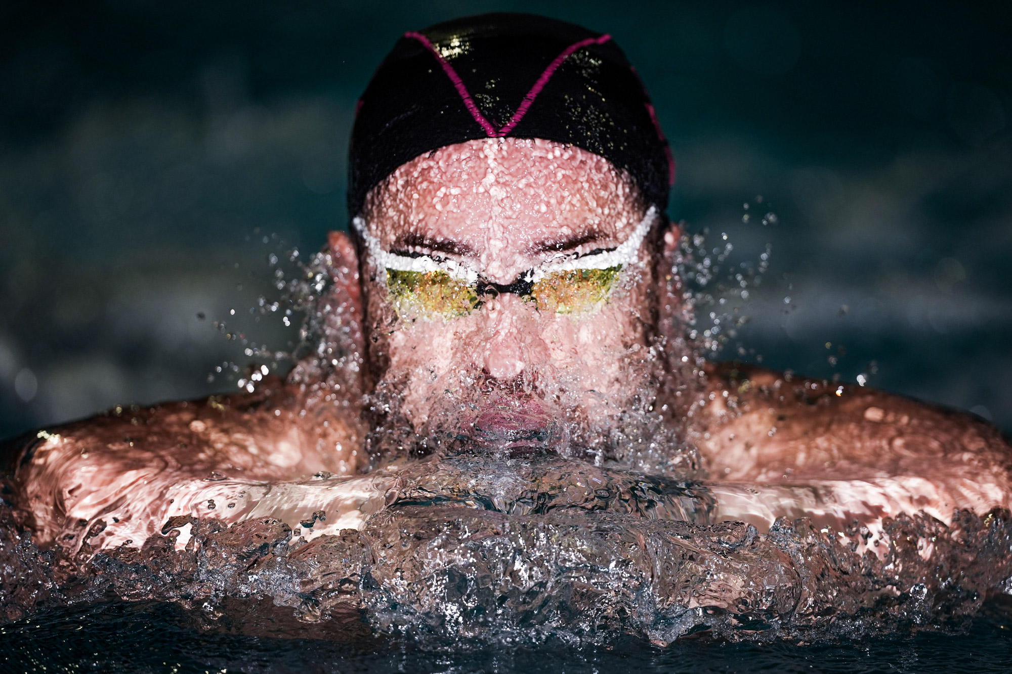 A swimmer wearing goggles and a swim cap rising out of the water still covered in droplets shot from directly in front.