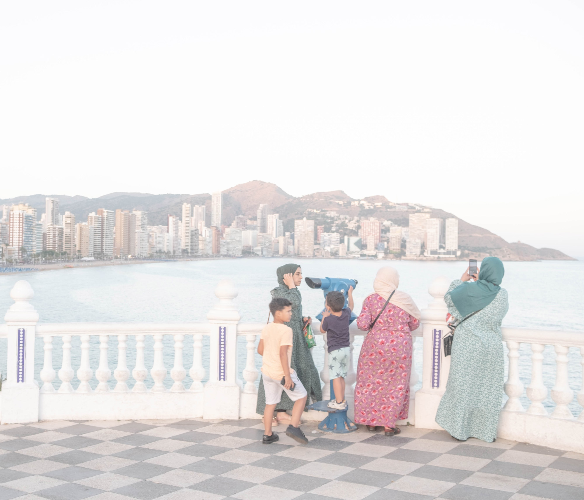 Five people standing near a telescope by some white railings overlooking a coastal city shot by Jordi Jon Pardo with a Fujifilm X-Pro 2