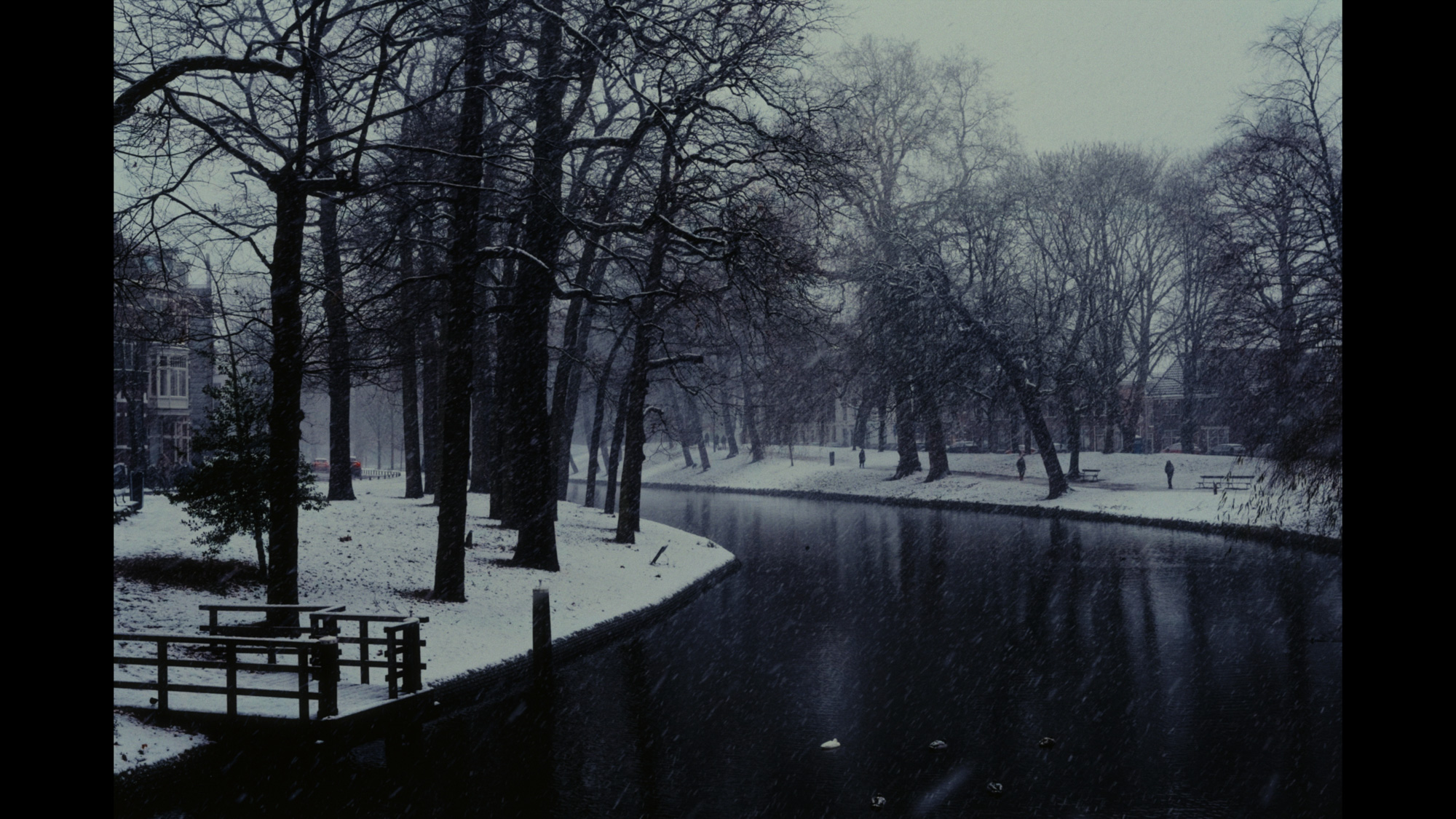 A tranquil canal meanders through a snow-covered park, lined with bare trees and a small wooden dock in the foreground.