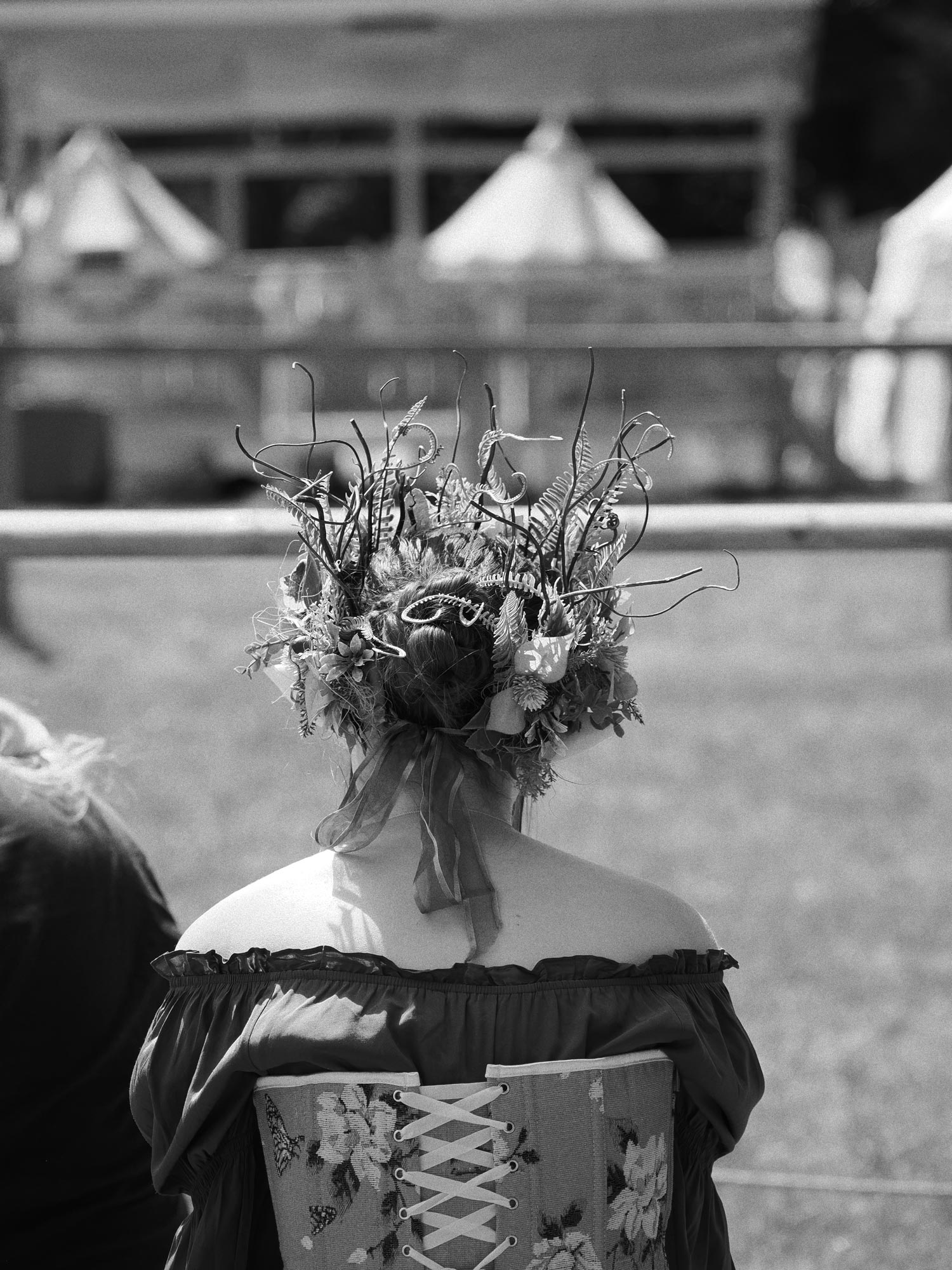A black and white photo of the back of a spectator at a medieval festival. The person is wearing a floral corset and has an intricate floral accessory on their head. 