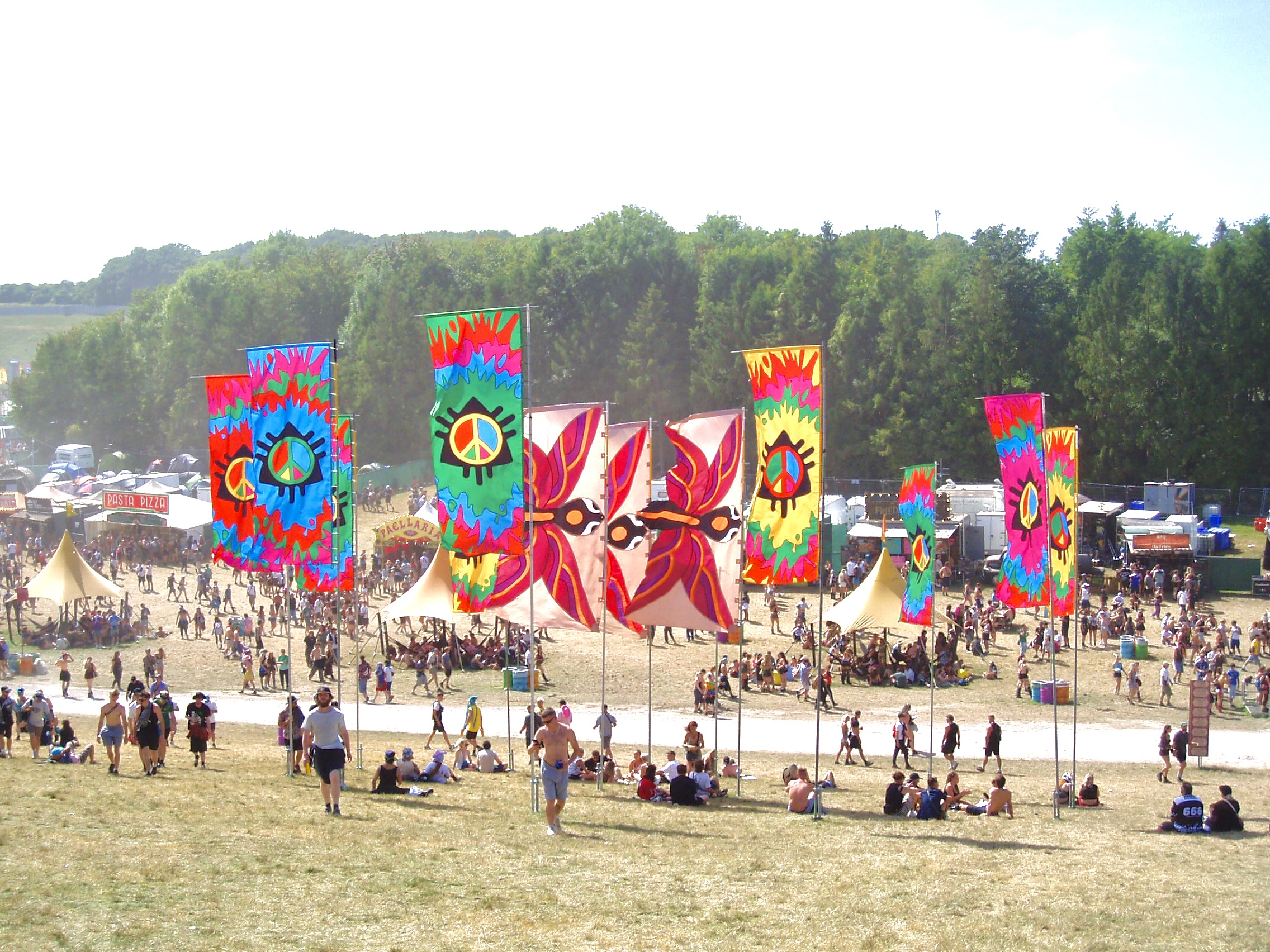 Uma coleção de grandes bandeiras coloridas num festival de música, com muitas pessoas a circular e sentadas pelo recinto.
