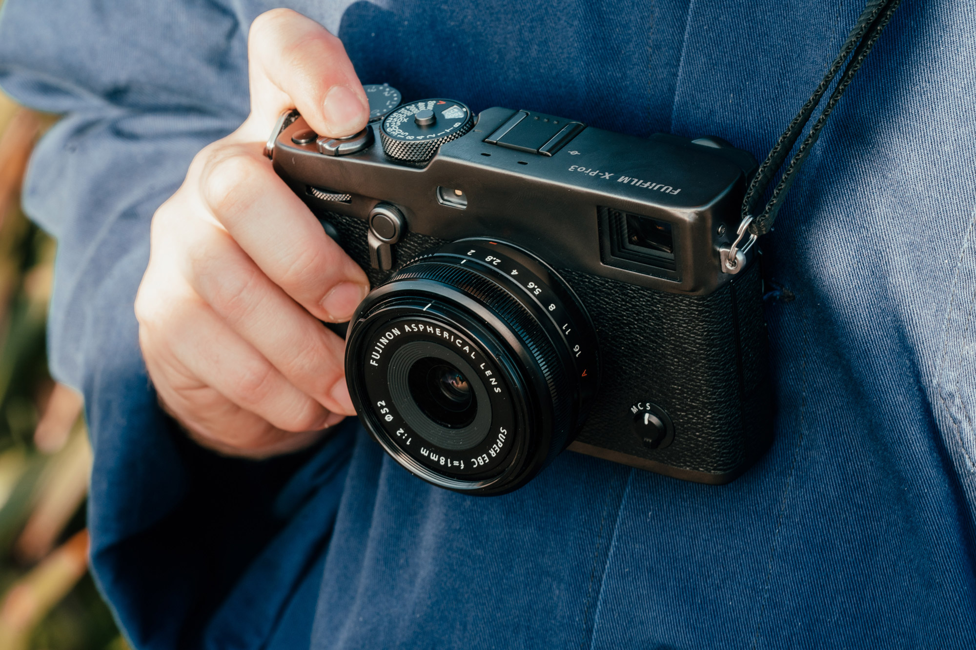 Gros plan d'un photographe portant une chemise bleue tenant un Fujifilm X-Pro 3 avec son doigt sur le déclencheur.