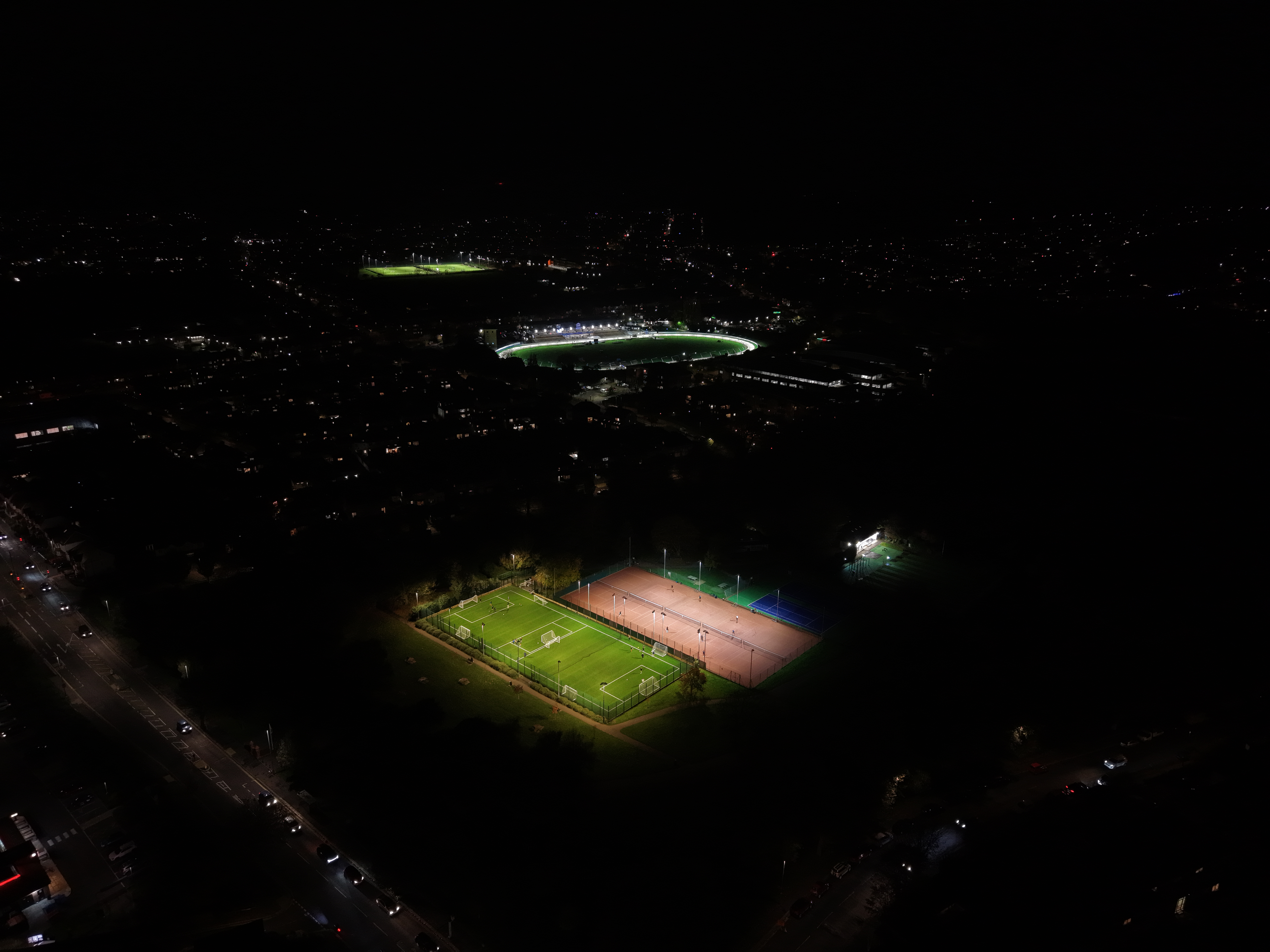 Imagem aérea noturna captada com o DJI Mini 5 Pro, mostrando campos de futebol e ténis iluminados numa zona urbana. A cena destaca o desempenho do drone em condições de pouca luz.