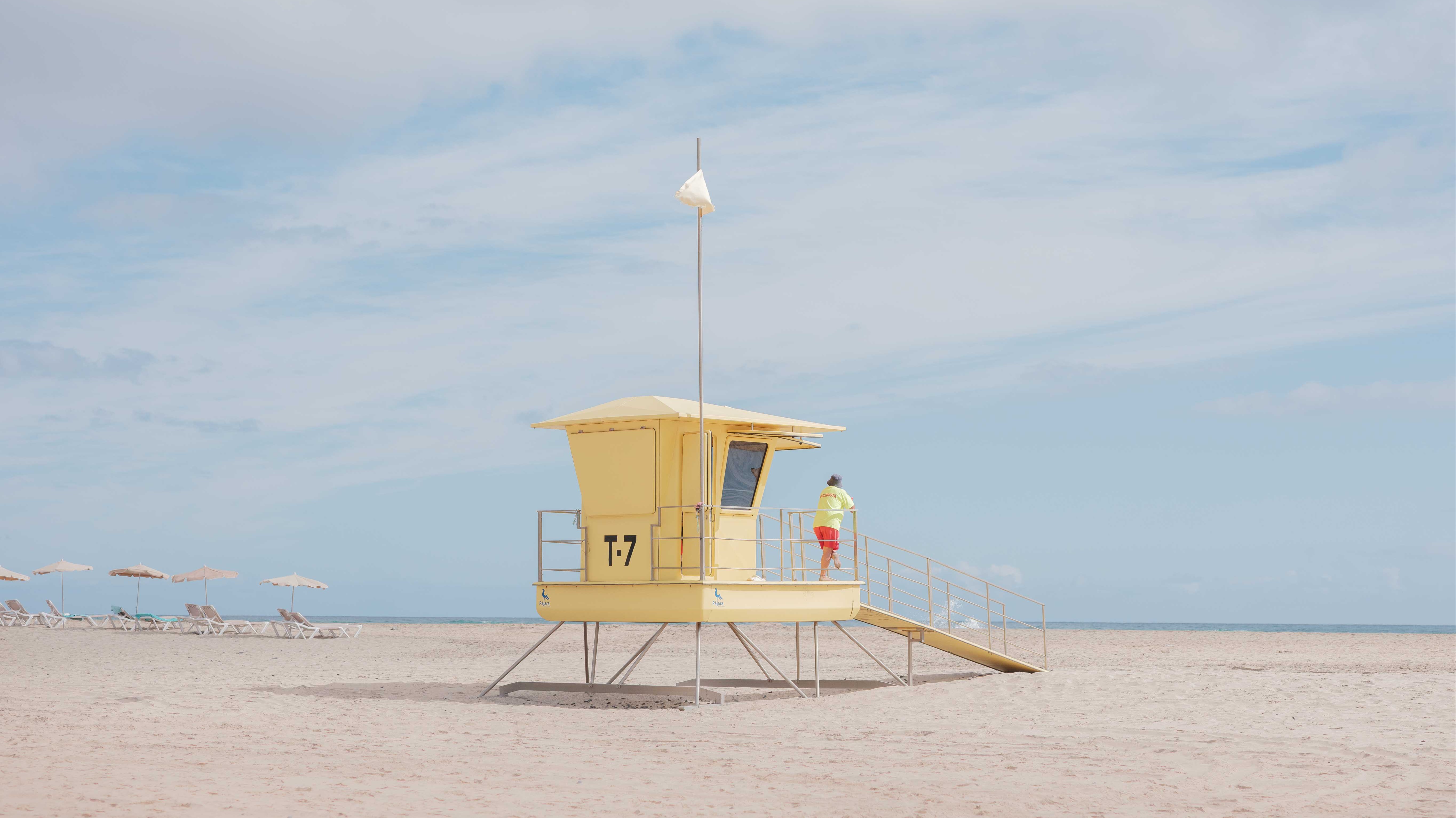 A yellow lifeguard hut on the beach with a lifeguard looking out to sea. By Connor Redmond.

